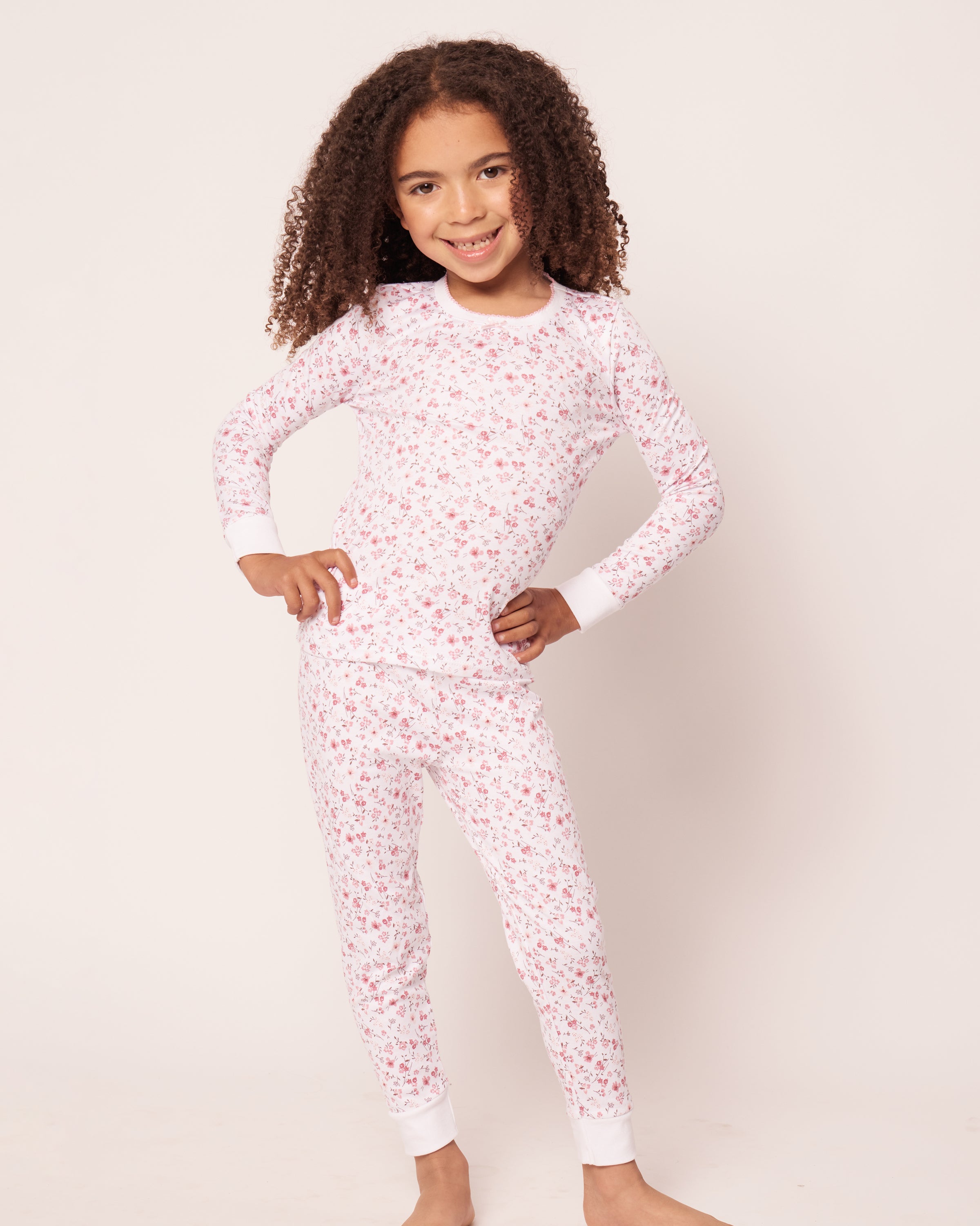 A curly-haired child beams, wearing Petite Plumes Kids Pima Snug Fit Pajama Set in Dorset Floral. The comfy white sleepwear set with a pink floral pattern features a long sleeve top and pants as they stand barefoot against a plain white background, hands on hips and feet slightly apart.
