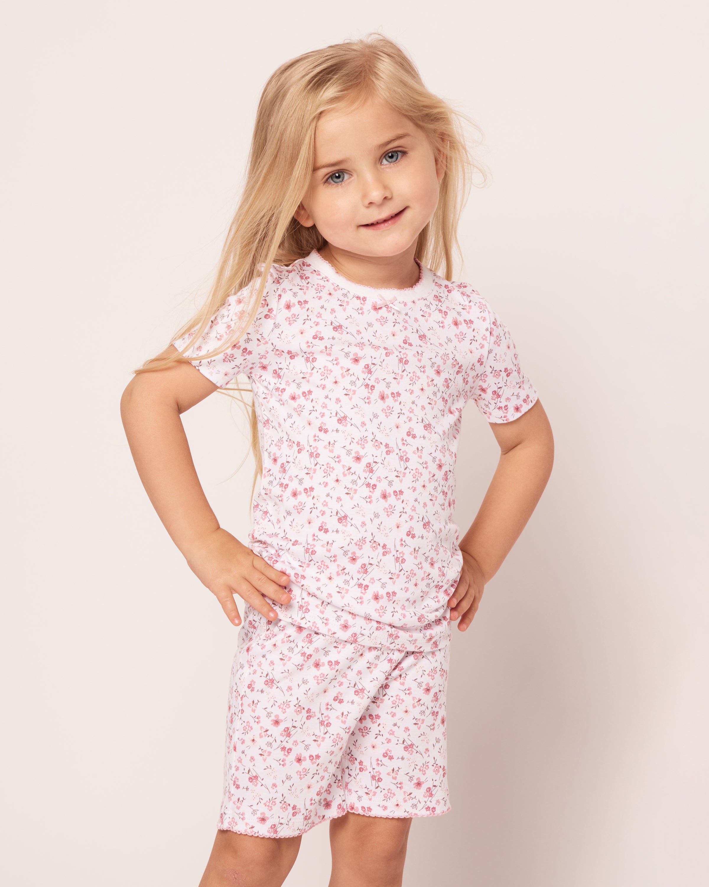 A young girl with long blonde hair wearing Petite Plumes Kids Pima Snug Fit Pajama Short Set in Dorset Floral stands against a plain background. Her hands on her hips, she smiles slightly, highlighting the comfort and style of the short sleeve top and shorts.