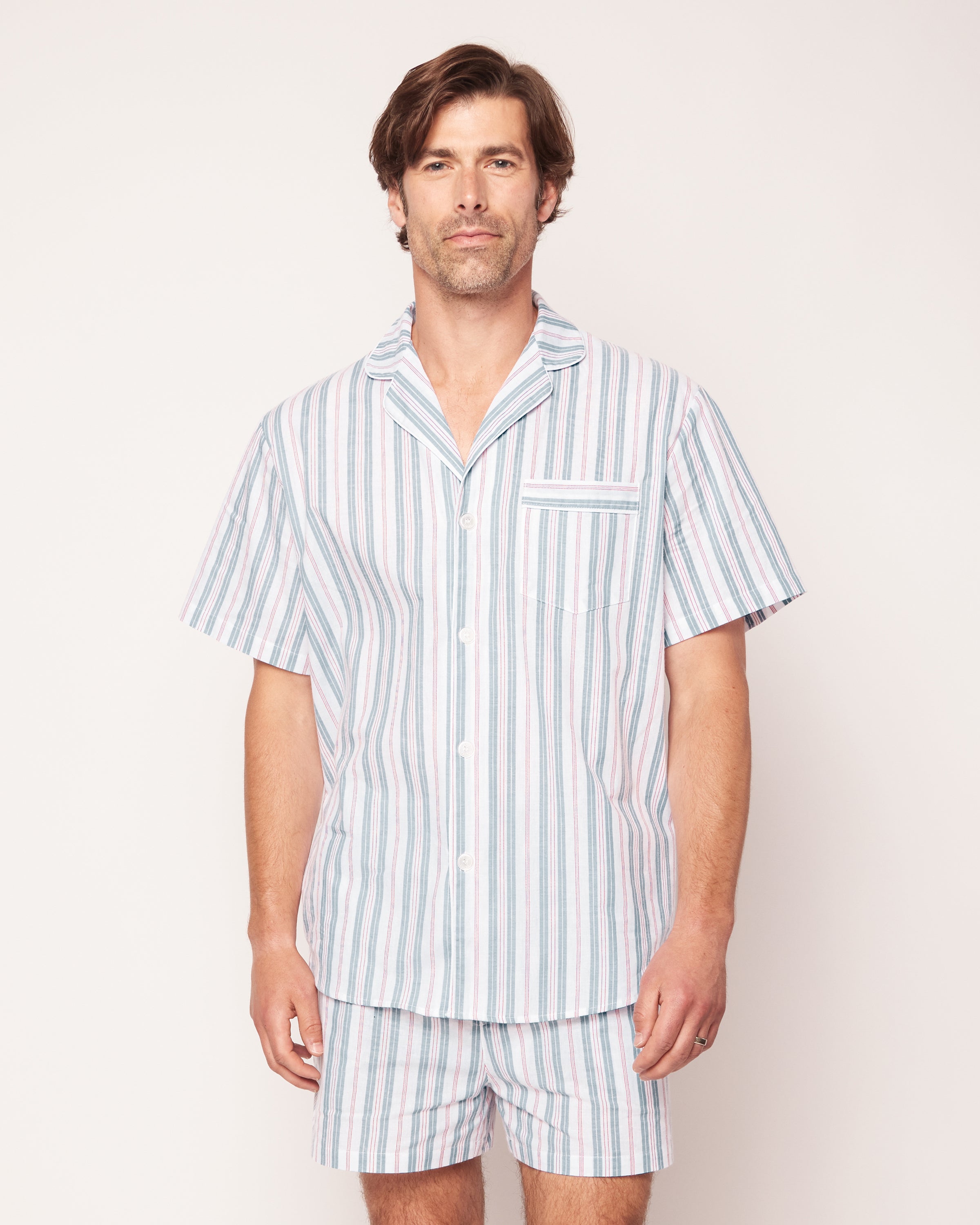 A person wearing Petite Plumes Mens Twill Pajama Short Set in Vintage French Stripes, featuring a vintage-style short-sleeved button-up shirt with collar and matching shorts in white, blue, and pink stripes, crafted from premium cotton against a light neutral background.