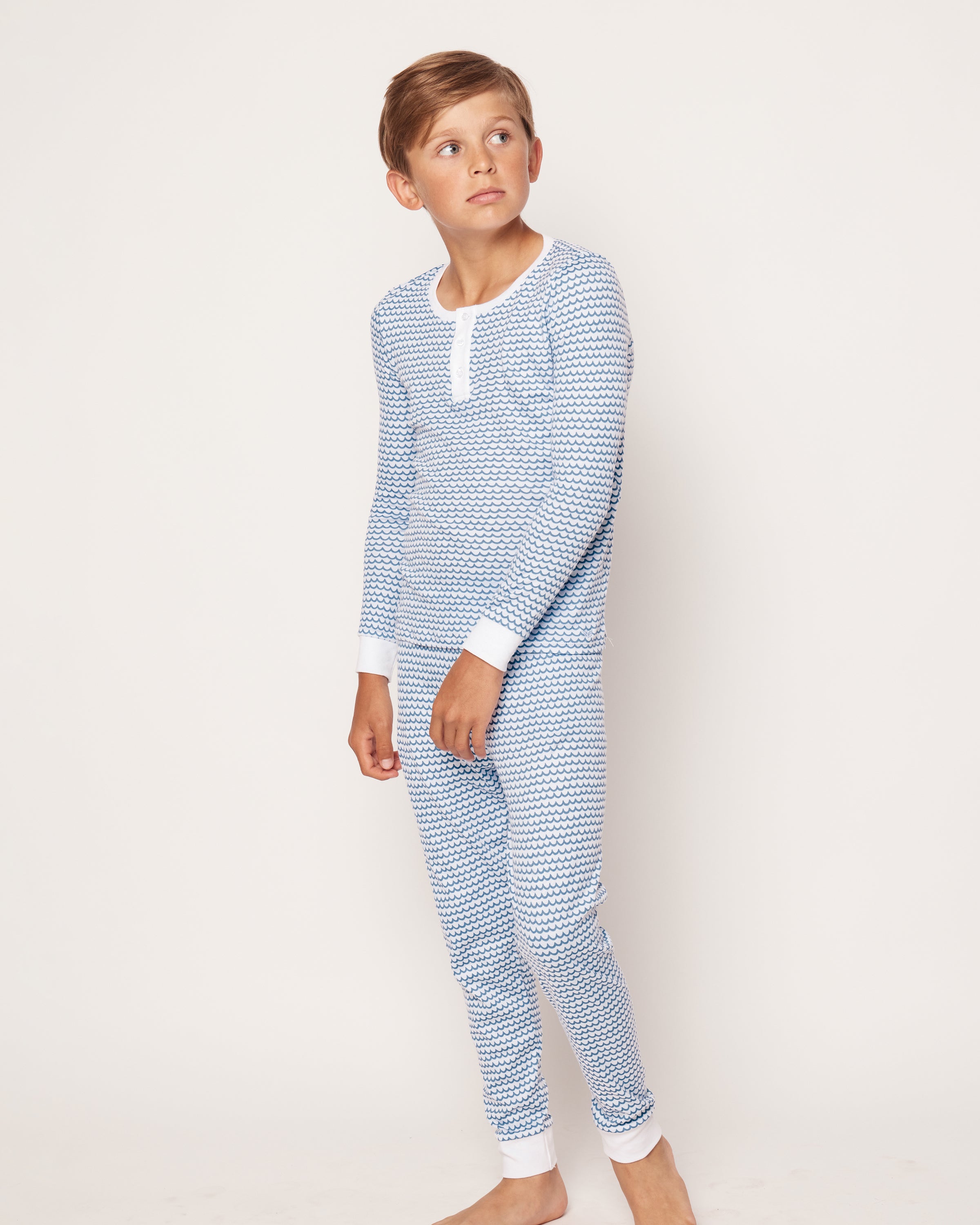 A young boy stands barefoot in a Petite Plume Kids Pima Snug Fit Pajama Set in La Mer, featuring a cozy long sleeve top and pant set crafted from soft Pima cotton with blue and white triangle patterns. The simple background emphasizes his short hair as he gazes thoughtfully to the side.