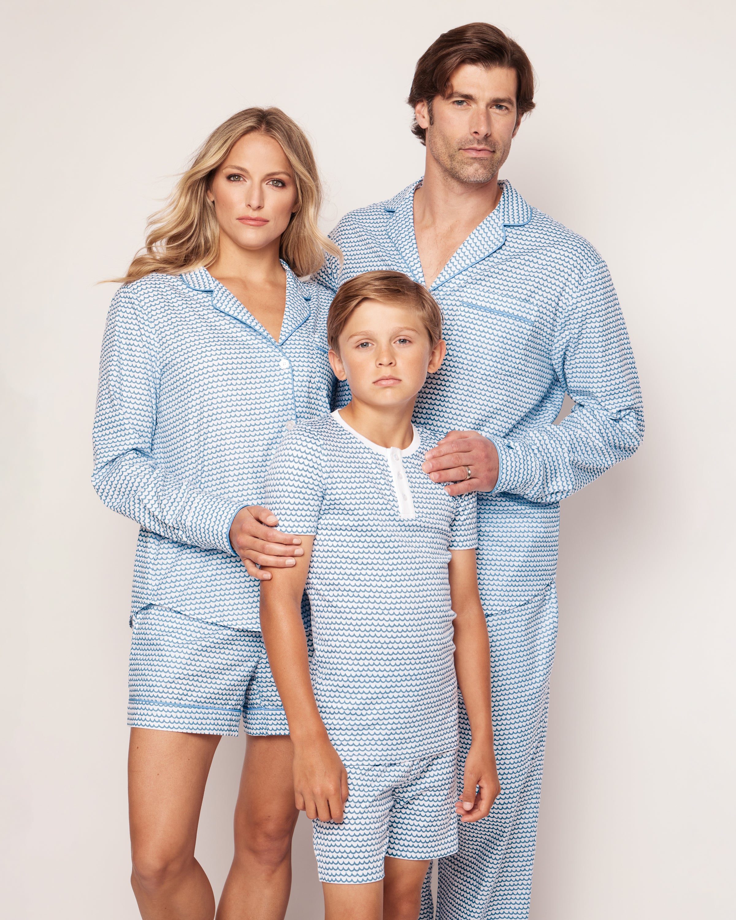 A family of three, clad in matching Petite Plumes Kids Pima Snug Fit Pajama Short Set in La Mer, stands against a white background. The parents, in cozy Pima cotton short sleeves, embrace their child lovingly.
