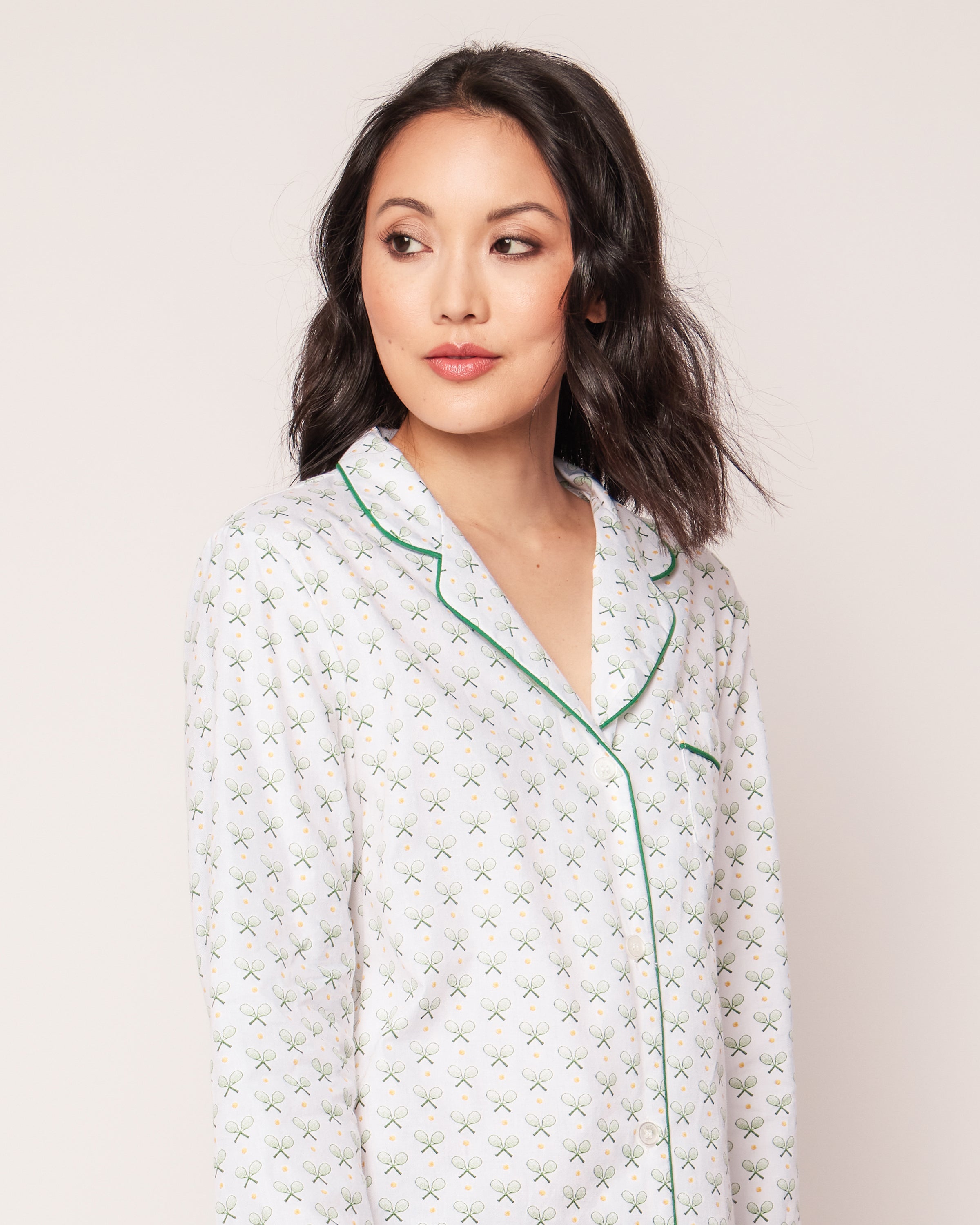 A person with shoulder-length dark hair is wearing the Womens Twill Nightshirt in Match Point from Petite Plume, showcasing luxury sleepwear. The pajama top is white with green trim and small green cross patterns as they look off to the side against a plain background.