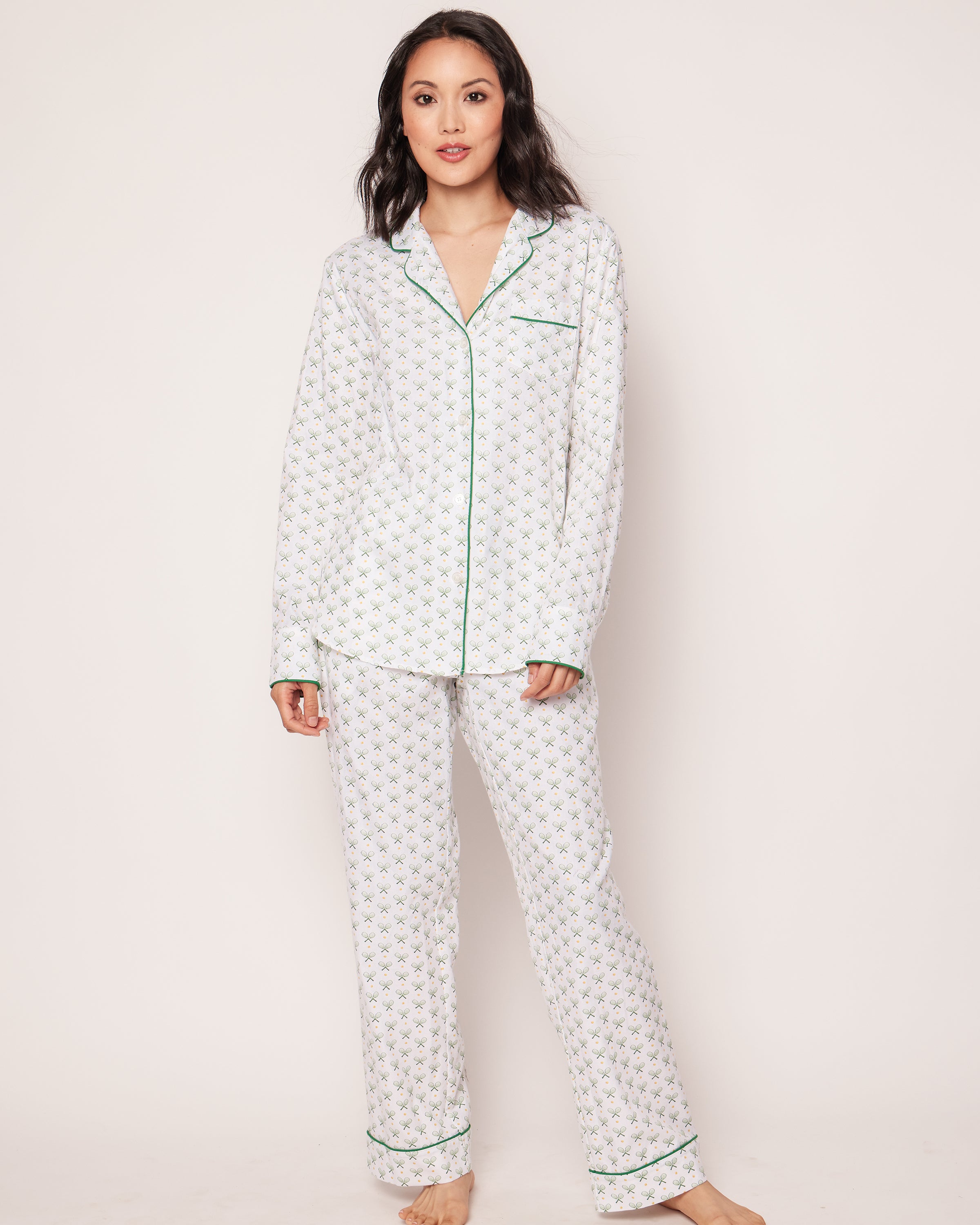 A person wears the Womens Twill Pajama Set in Match Point by Petite Plume, made from premium cotton with a white floral pattern and green trim. The set includes a long-sleeve top and pants, perfect for cozy relaxation, as they stand barefoot against a plain background.