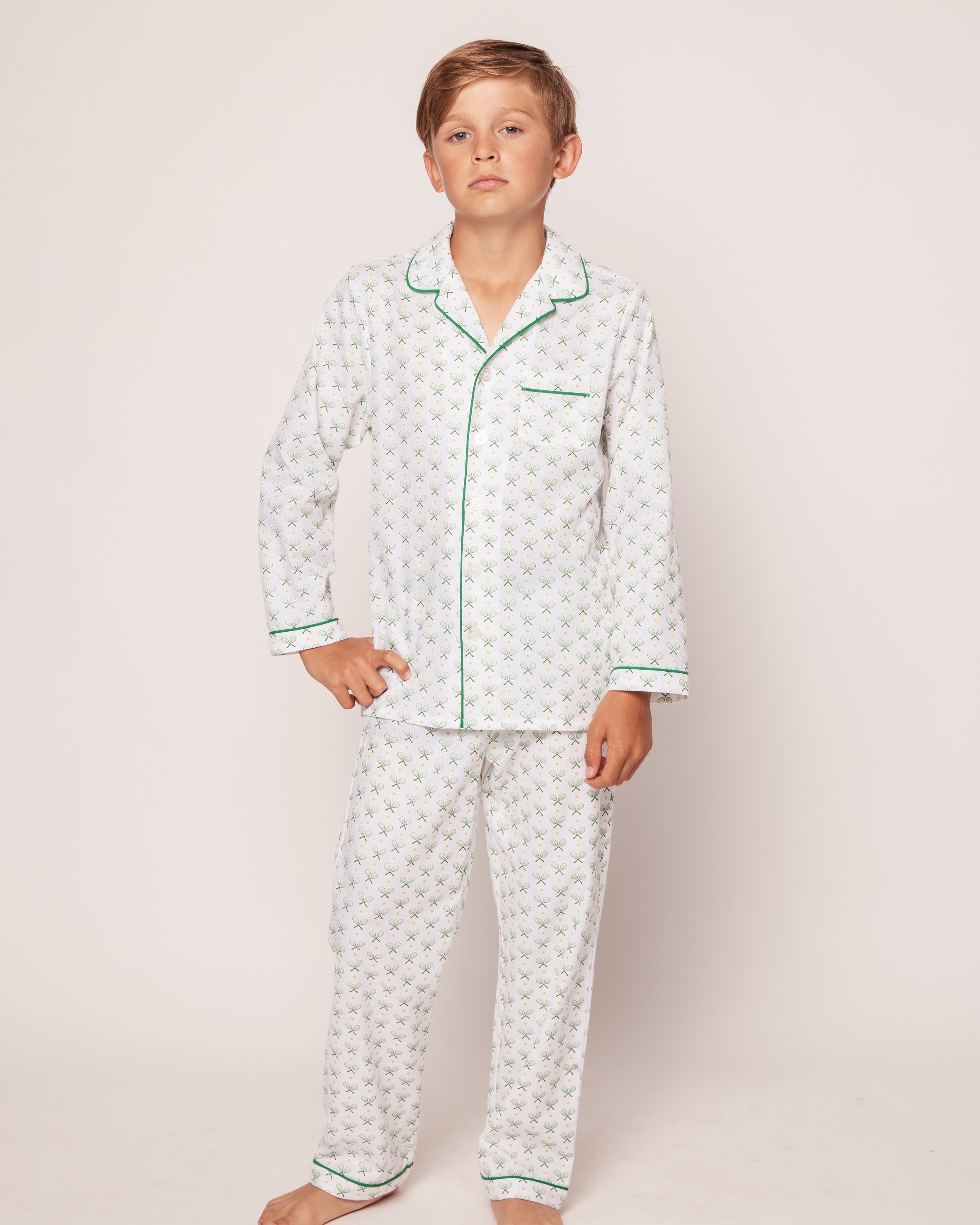 A young boy stands barefoot in Petite Plumes Kids Twill Pajama Set in Match Point, featuring white pajamas with a green floral pattern and trim. His light brown hair and relaxed expression complement the classic set against a plain, light background.
