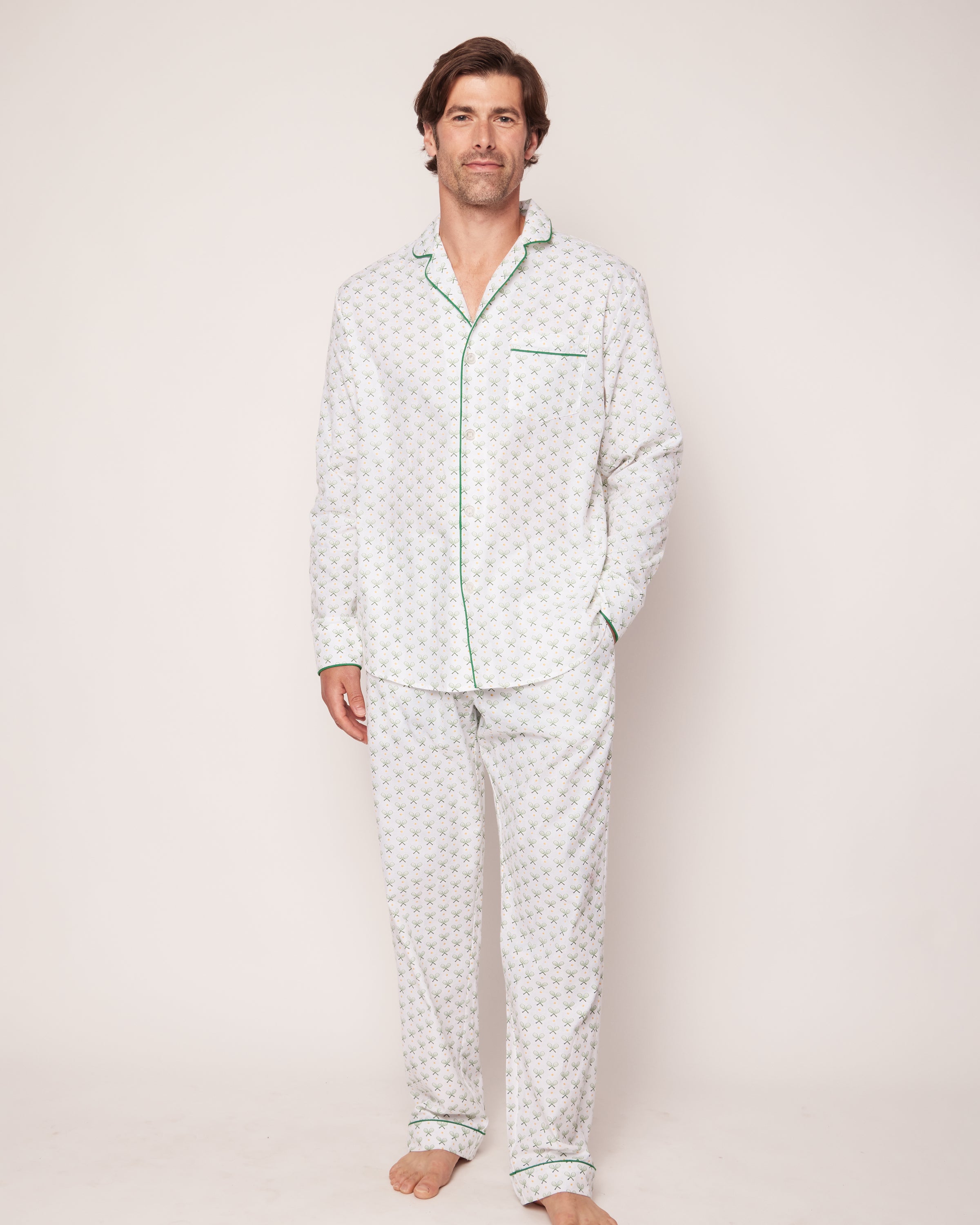A man stands barefoot on a plain background, wearing Petite Plumes Mens Twill Pajama Set in Match Point. The set features a tennis print with green piping and is crafted from high-quality cotton, including a long-sleeve top and pants. He faces forward in a casual pose.