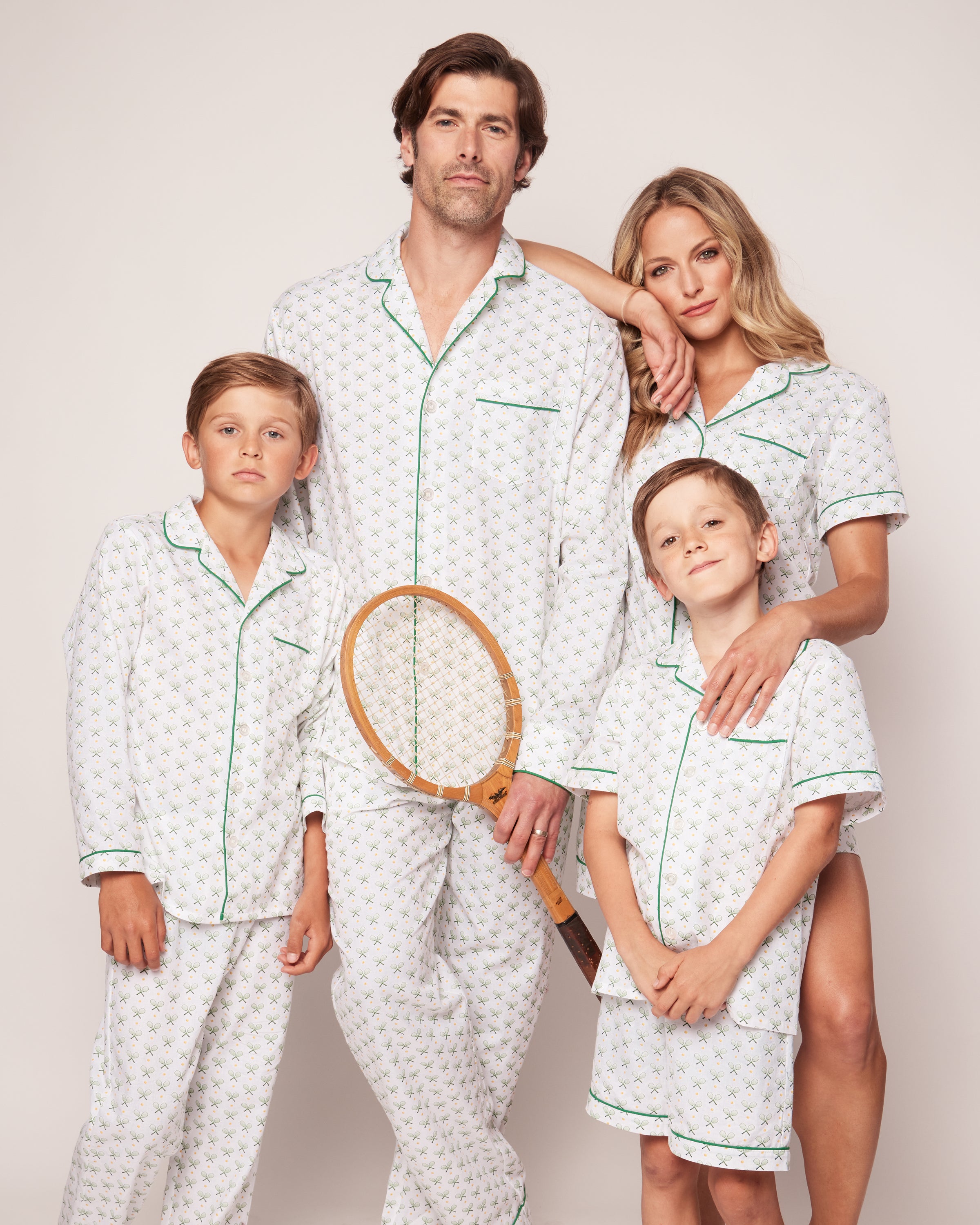 A family of four, wearing Petite Plumes Mens Twill Pajama Set in Match Point, stands together. The father holds a tennis racket beside the mother while two boys pose confidently. Their white pajamas with green piping perfectly blend comfort and style against a plain background.