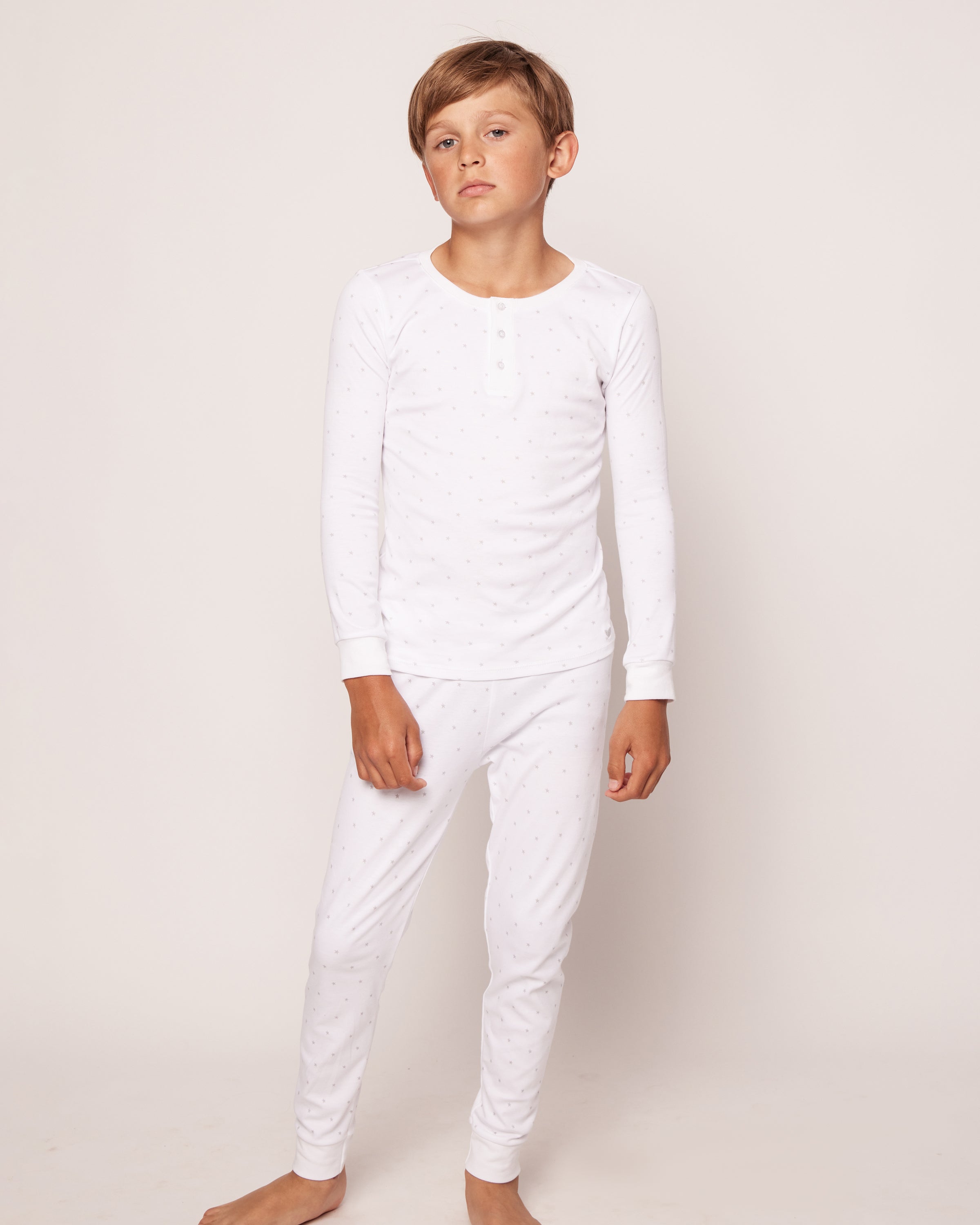 A young boy stands barefoot against a plain background, wearing the Kids Pima Snug Fit Pajama Set in Grey Stars from Petite Plume. Crafted from soft Pima Cotton, he exudes calmness and relaxation, perfect for showcasing quality sleepwear comfort.