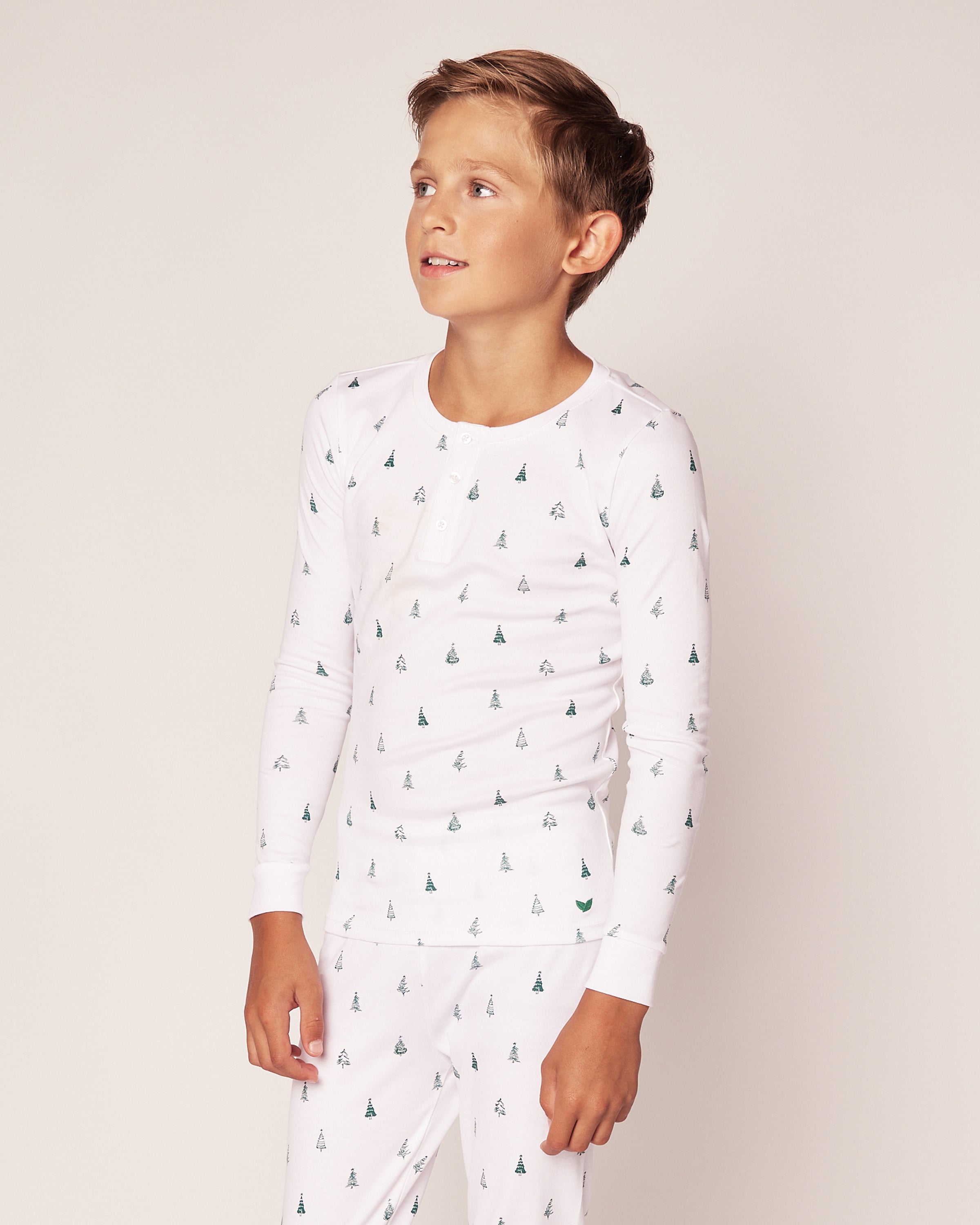 A child in Petite Plumes Kids Pima Snug Fit Pajama Set in Evergreen Trees, made of soft Pima cotton, stands against a plain background looking to the side. The long sleeve top and pant set exudes a light and neutral tone.