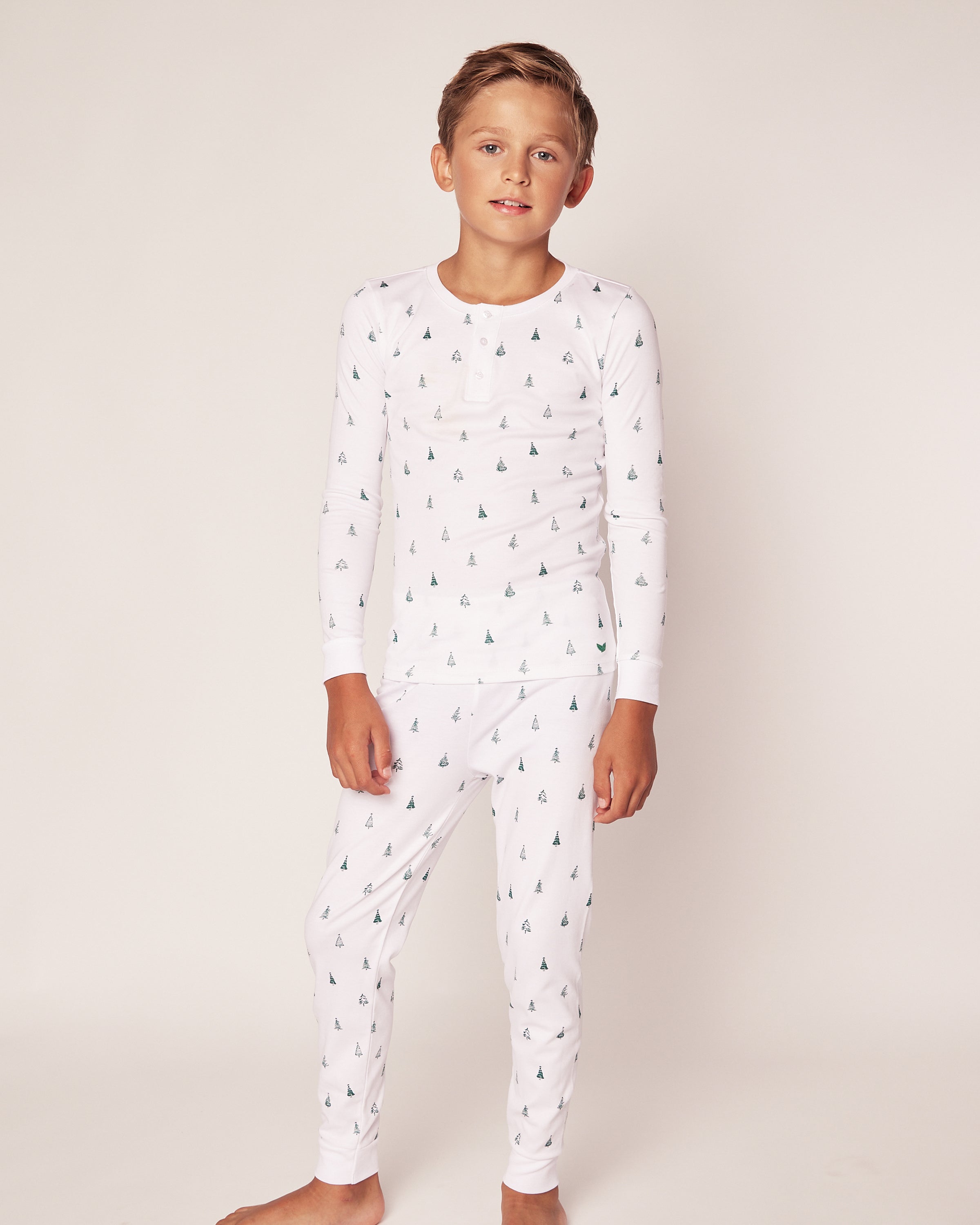 A child with short, light brown hair smiles slightly while standing barefoot in a Petite Plume Kids Pima Snug Fit Pajama Set in Evergreen Trees. The long sleeve top and pant set has small tree patterns on soft Pima cotton. They stand against a plain, light-colored background with hands by their sides.