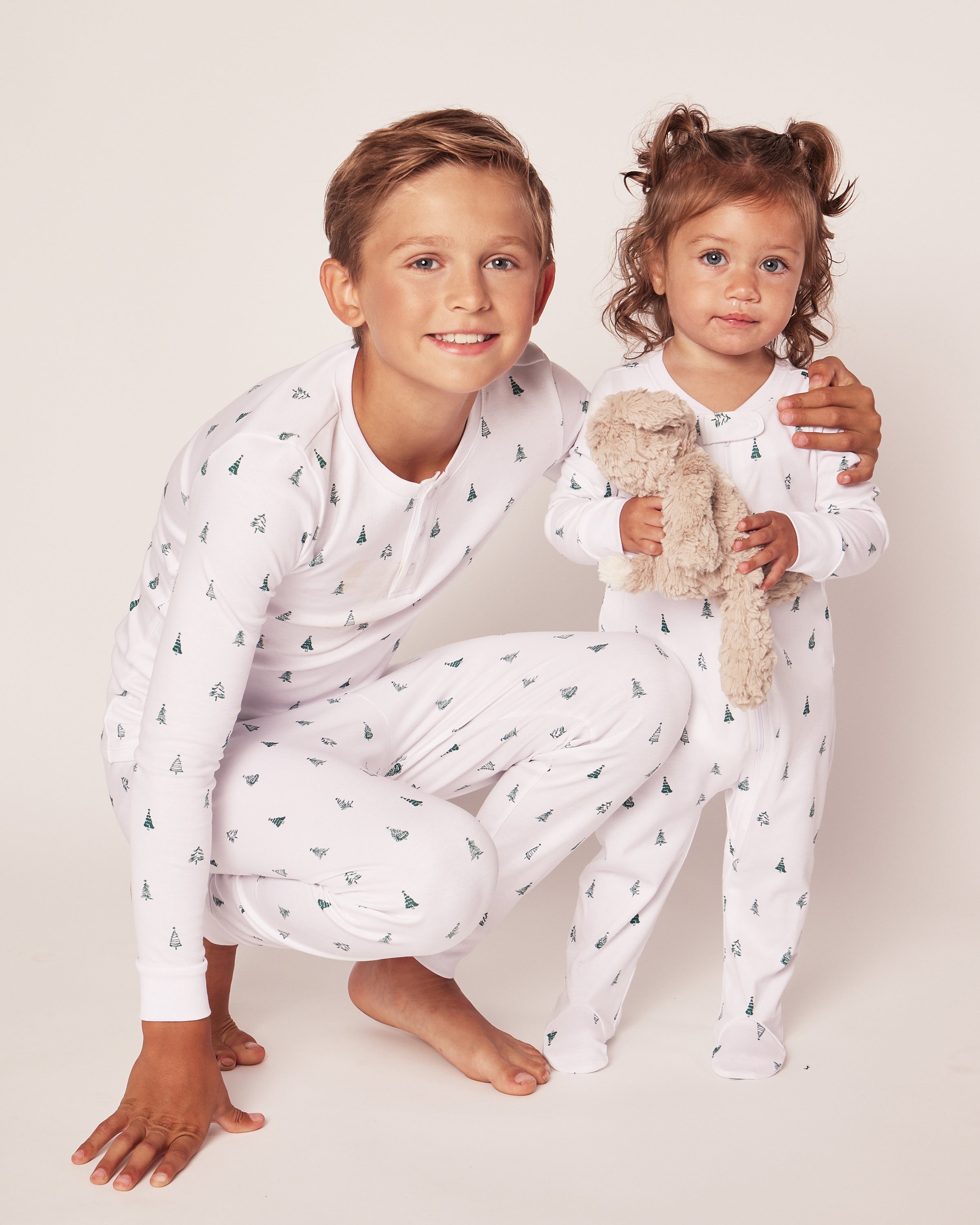 Two smiling children are photographed wearing Petite Plumes Kids Pima Snug Fit Pajama Set in Evergreen Trees. The older child wears a long sleeve top and pants while crouched beside the younger one holding a plush toy, set against a plain, light-colored background.