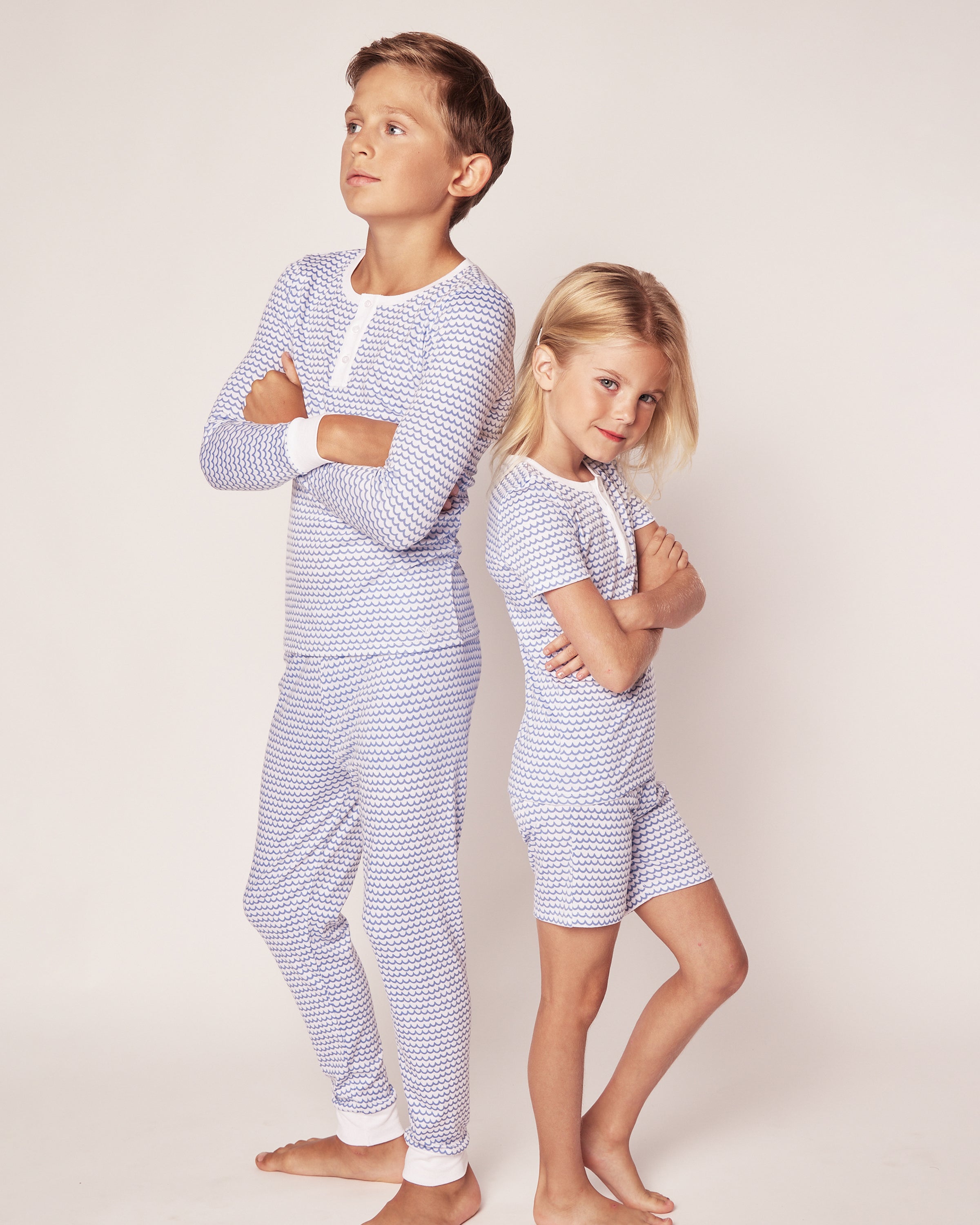 Two children stand confidently back-to-back, arms crossed, in matching Petite Plume Kids Pima Snug Fit Pajama Sets in La Mer. Crafted from soft Pima cotton, the sleepwears blue and white pattern pops against a plain background, highlighting their posture and expressions.