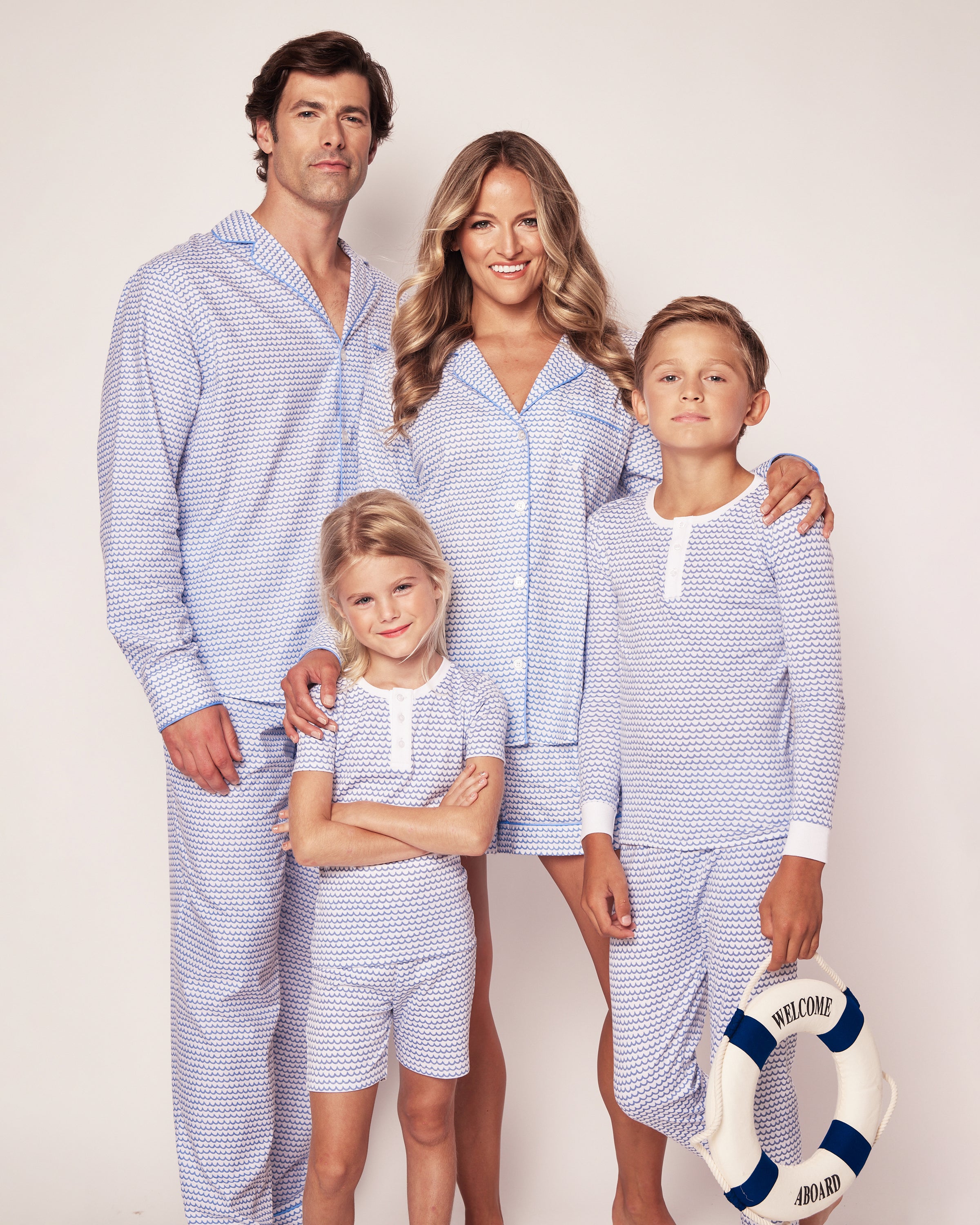 A family of four stands together in matching Petite Plumes Kids Pima Snug Fit Pajama Set in La Mer. The boy holds a life preserver with WELCOME ABOARD. Their smiles shine against the plain background as they cozy up in their long sleeve top and pant set.