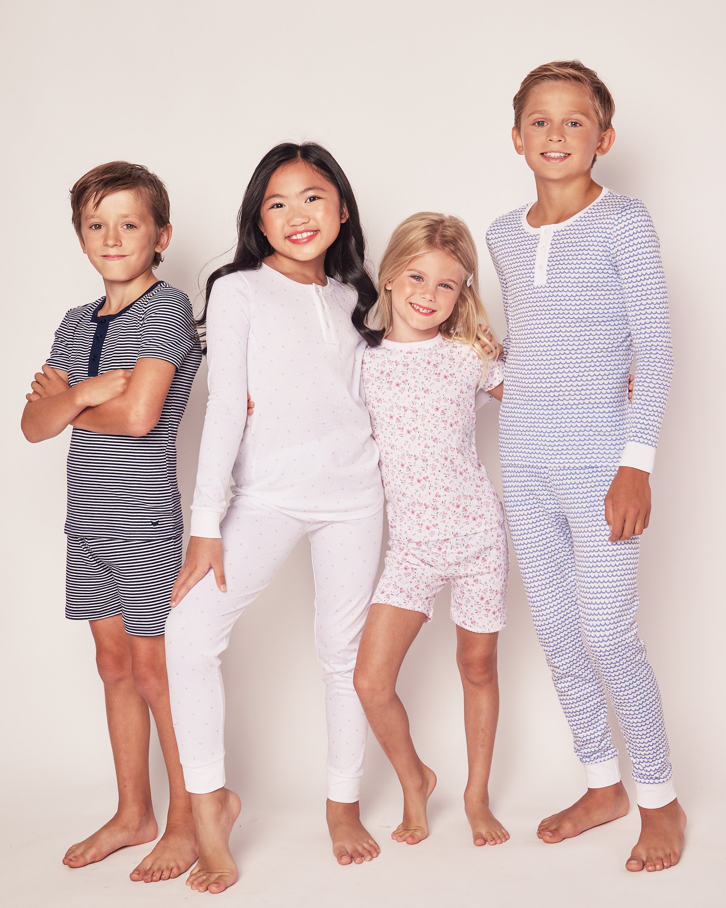 Four smiling children casually pose against a plain background in coordinated styles, wearing Petite Plumes Kids Pima Snug Fit Pajama Set in Grey Stars. The group of two boys and two girls showcases soft-colored, patterned tops and pants with stripes, dots, and floral designs.