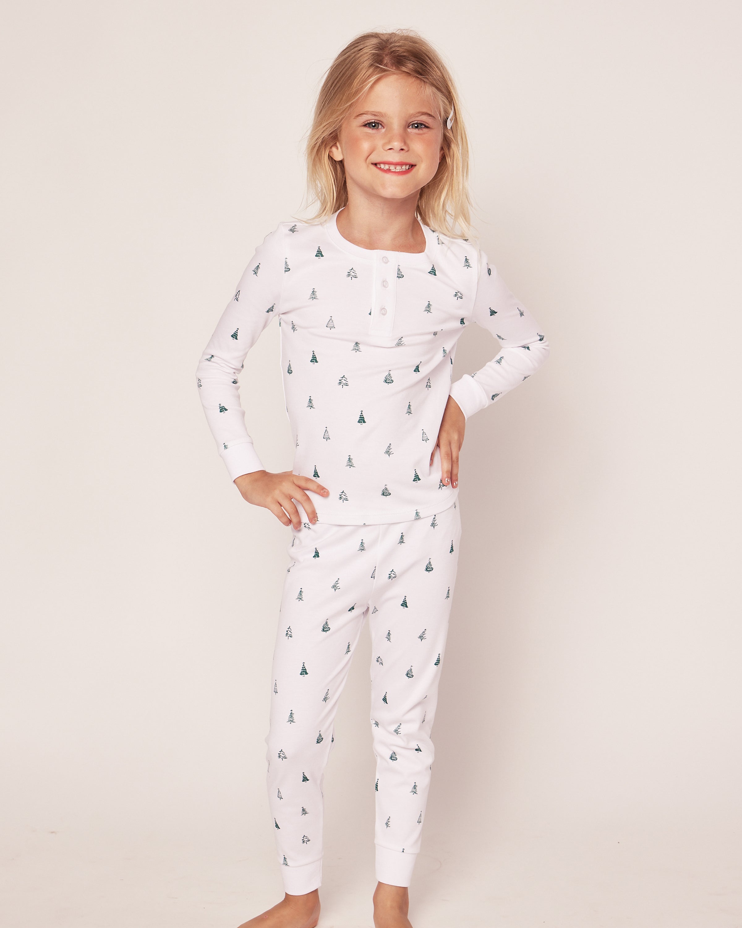 A smiling child with long blonde hair models Petite Plumes Kids Pima Snug Fit Pajama Set in Evergreen Trees, confidently posing with hands on hips against a plain backdrop in their cozy, luxurious long sleeve top and pants made of Pima cotton.