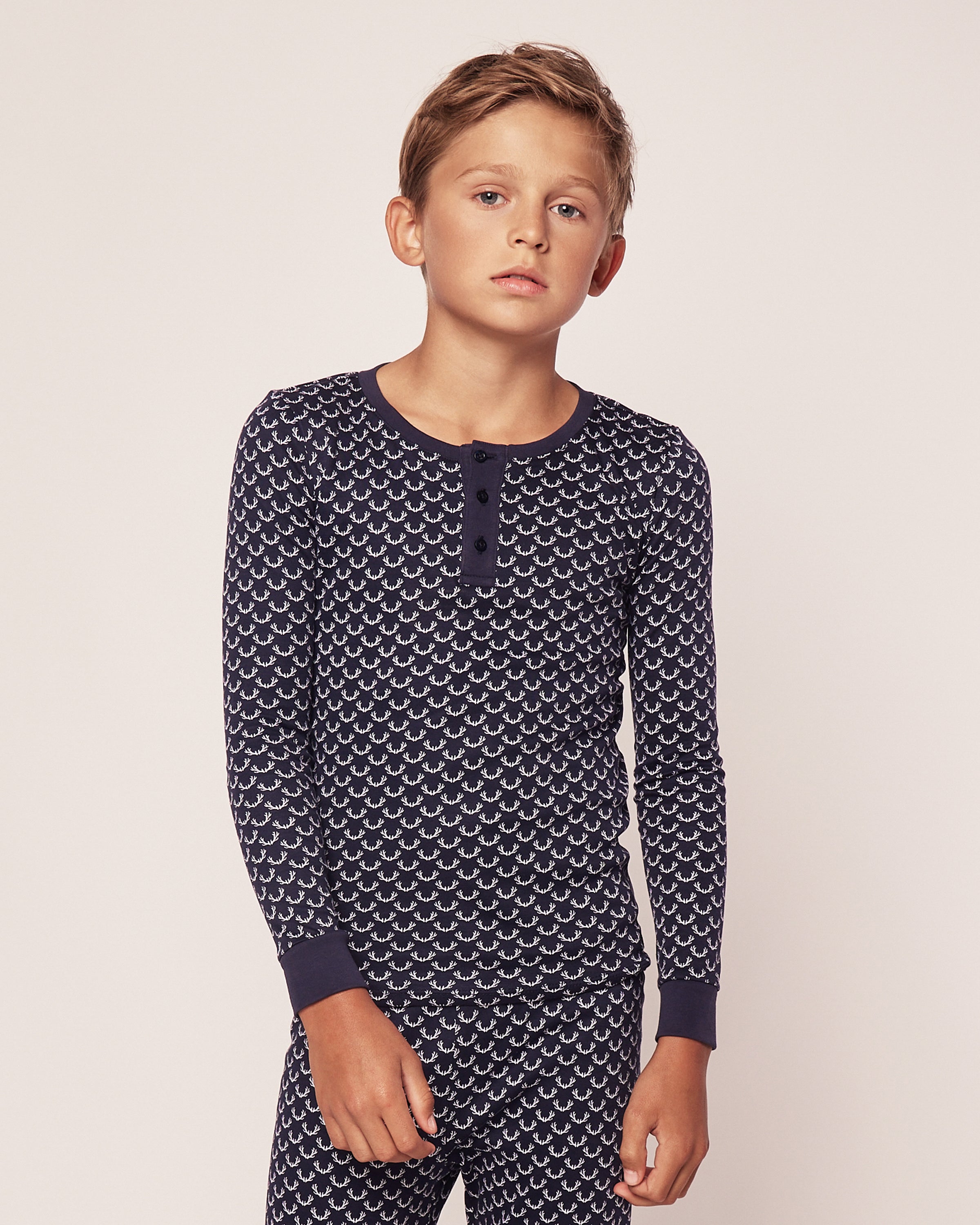 A young boy stands against a plain background wearing Petite Plumes Kids Pima Snug Fit Pajama Set Nordic Antlers, a navy blue long-sleeve top and pant set with white pattern. Made from Pima cotton, the shirt features a button-up collar. He looks at the camera with a neutral expression.