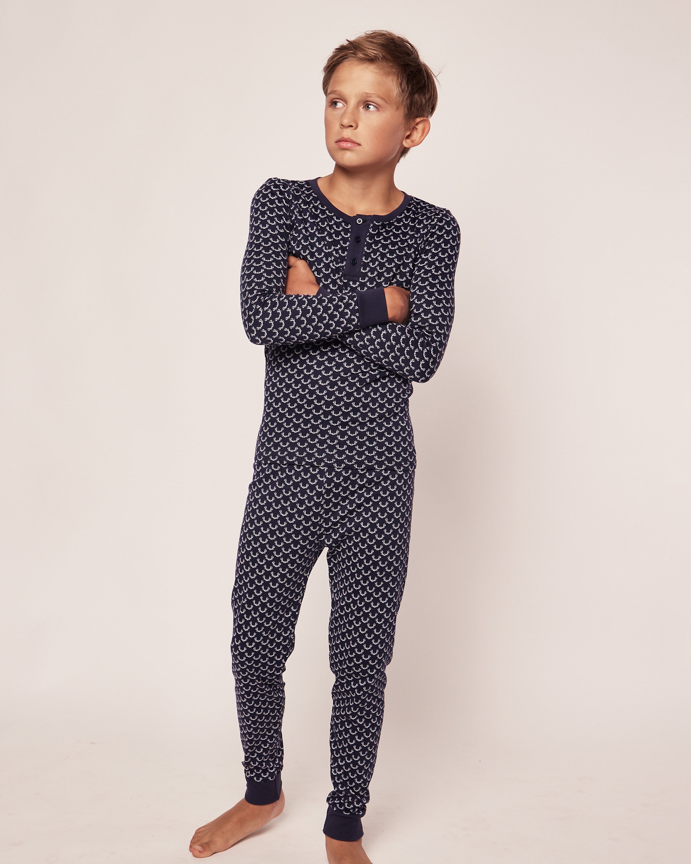 A young boy stands barefoot in a studio wearing the Petite Plume Kids Pima Snug Fit Pajama Set in Nordic Antlers. He crosses his arms and gazes sideways against a plain, light-colored backdrop, capturing a cozy sleepwear vibe.