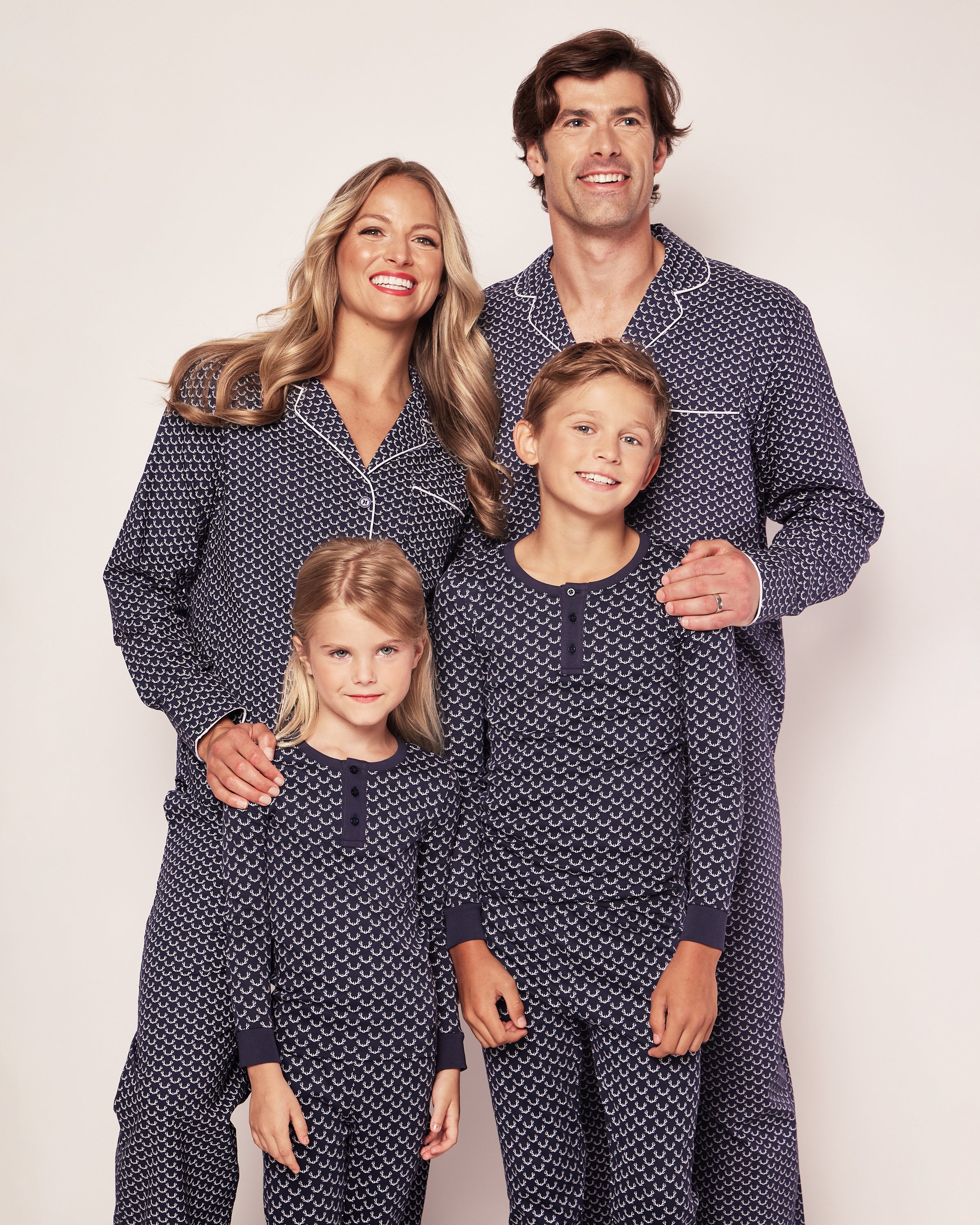 A family of four wears matching Petite Plumes Kids Pima Snug Fit Pajama Set in Nordic Antlers, with the adults behind and a child in front of each. The soft Pima cotton complements the plain white backdrop, emanating happiness and relaxation.