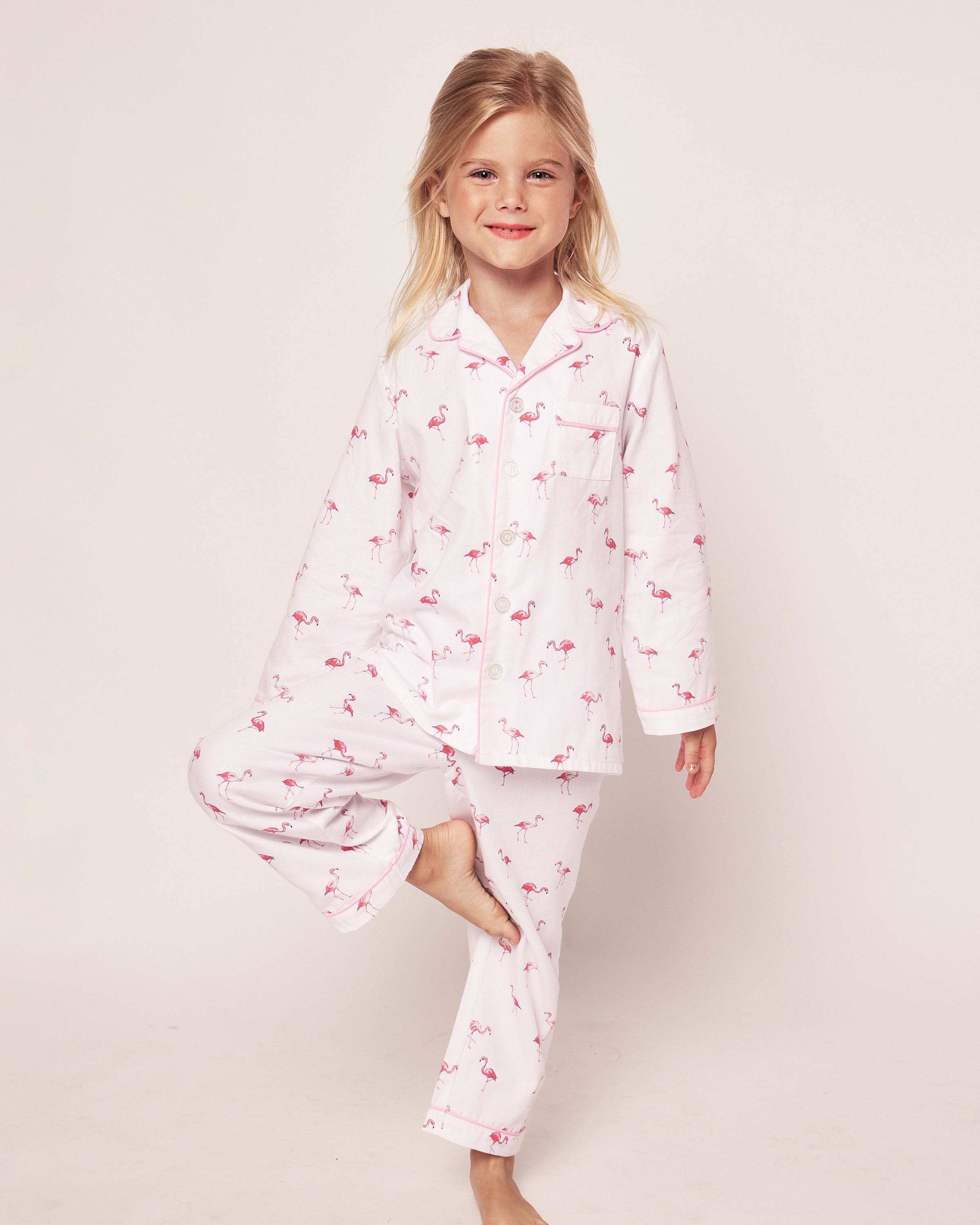 A young child with long blonde hair balances confidently on one foot, knee lifted, in a Petite Plume Kids Twill Pajama Set in Flamingos, featuring pink flamingo patterns. The flame retardant cotton outfit stands out against the plain white background as they smile and pose.