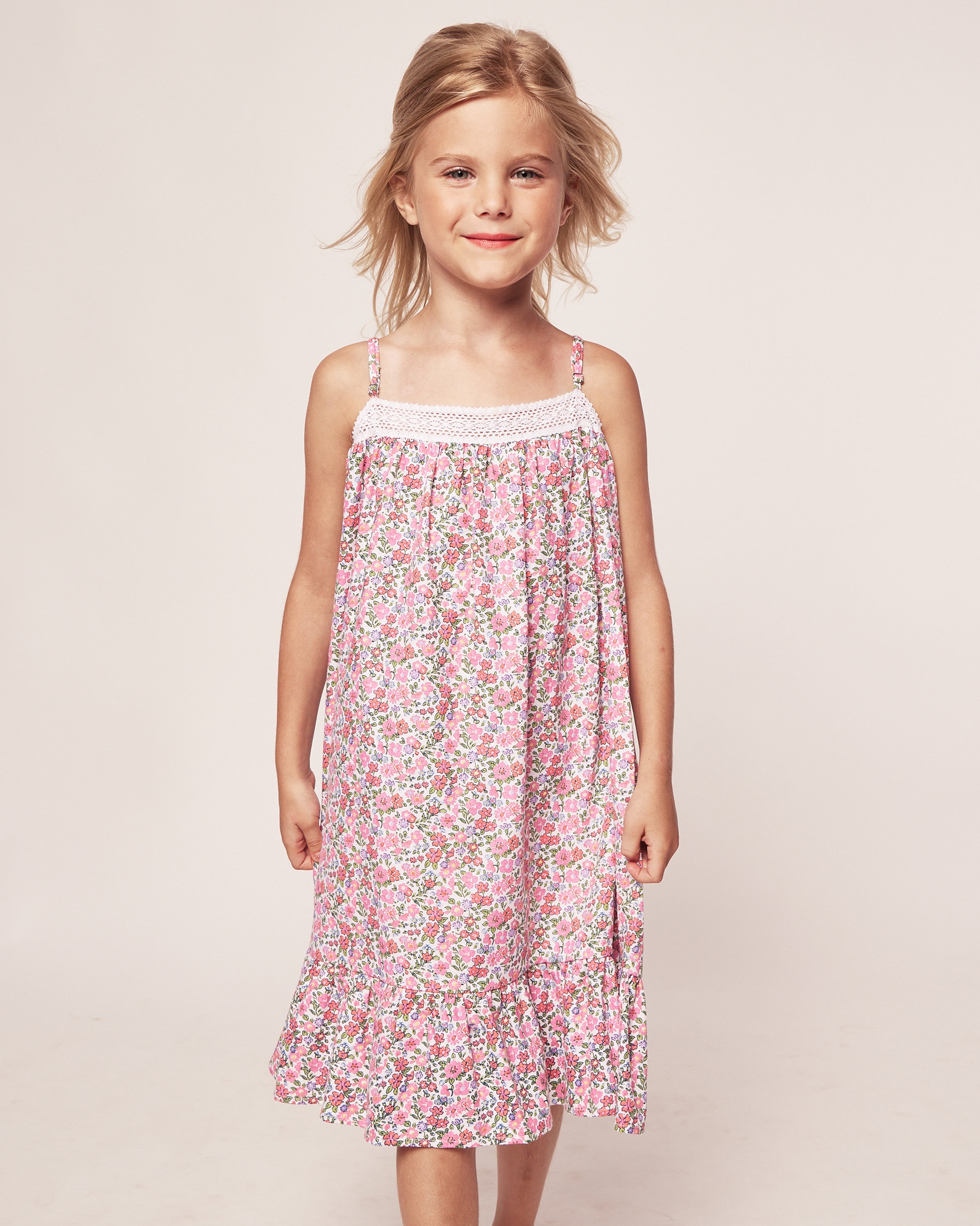 A young girl in Petite Plumes Girls Twill Lily Nightgown in Fleurs de Rose smiles while standing against a plain background. Her shoulder-length hair is parted to the side.