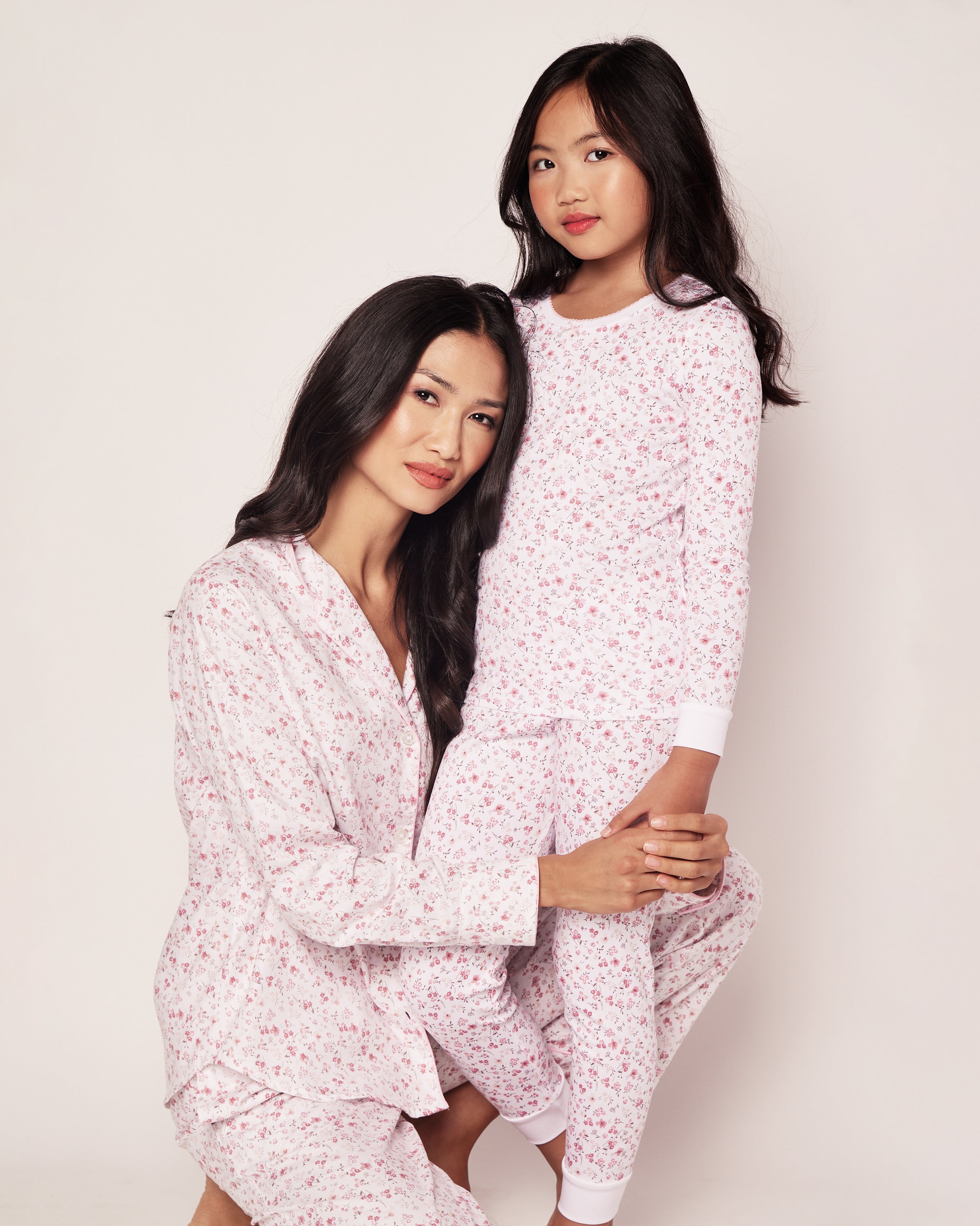 A woman and a girl stand against a plain background in matching Petite Plumes Dorset Floral pink sleepwear. The soft Pima Cotton long sleeve top and snug fit pant set keeps them cozy as the woman sits gracefully while the girl stands beside her, both smiling softly.