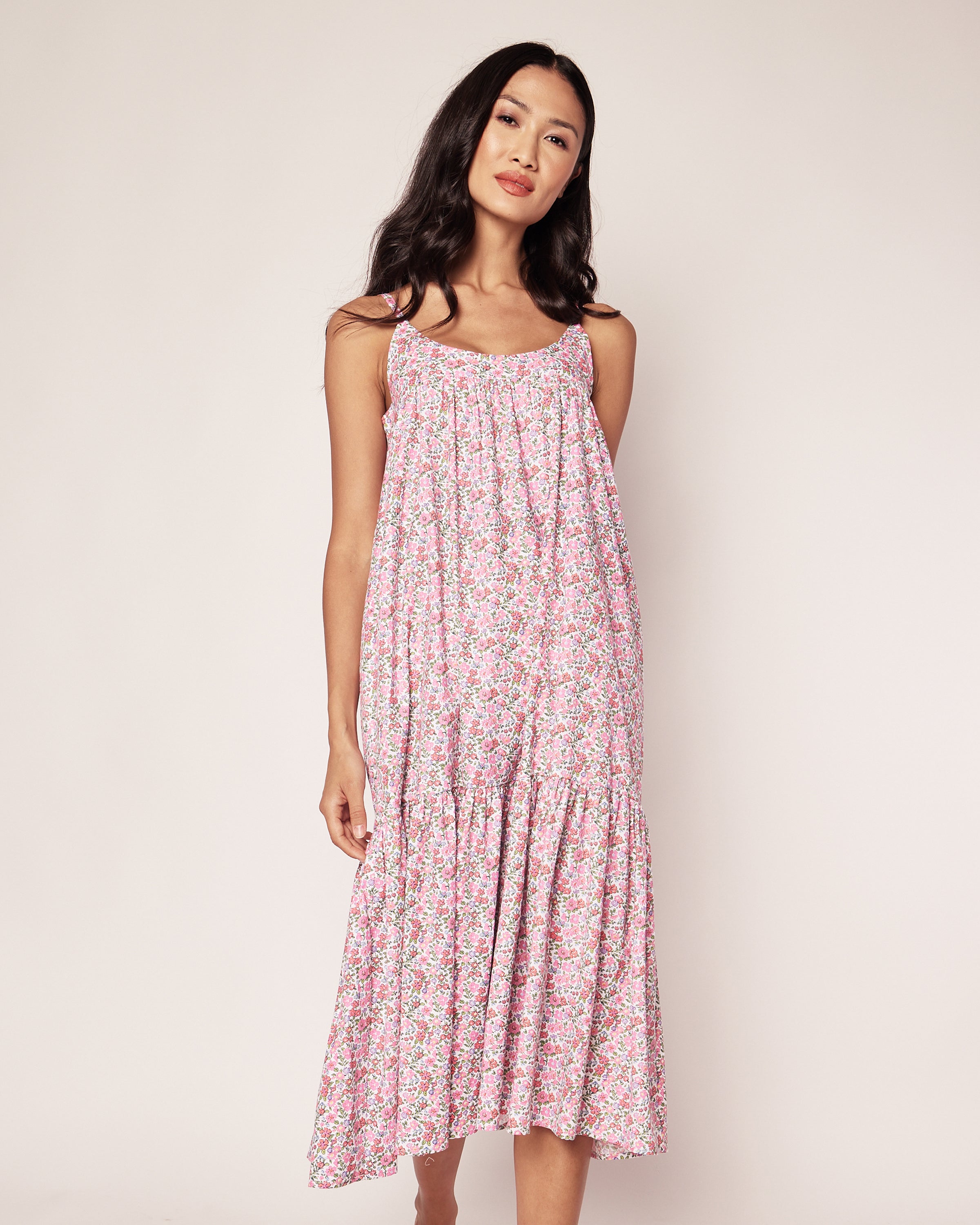 A person wearing Petite Plumes Womens Twill Chloe Nightgown in Fleurs de Rose stands against a plain light background. The flowy, ankle-grazing nightgown features a pink floral print. They have long, dark hair and a relaxed expression.