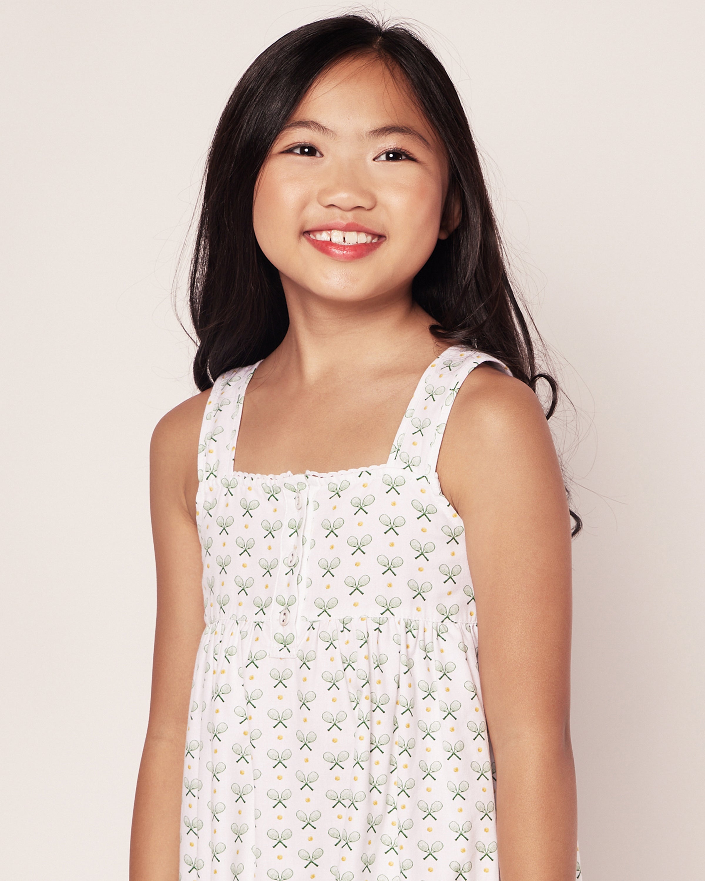 A young girl smiles, wearing the Petite Plume Girls Twill Charlotte Nightgown in Match Point. Her long black hair cascades over her shoulders, embodying comfort and style against a plain, light background.