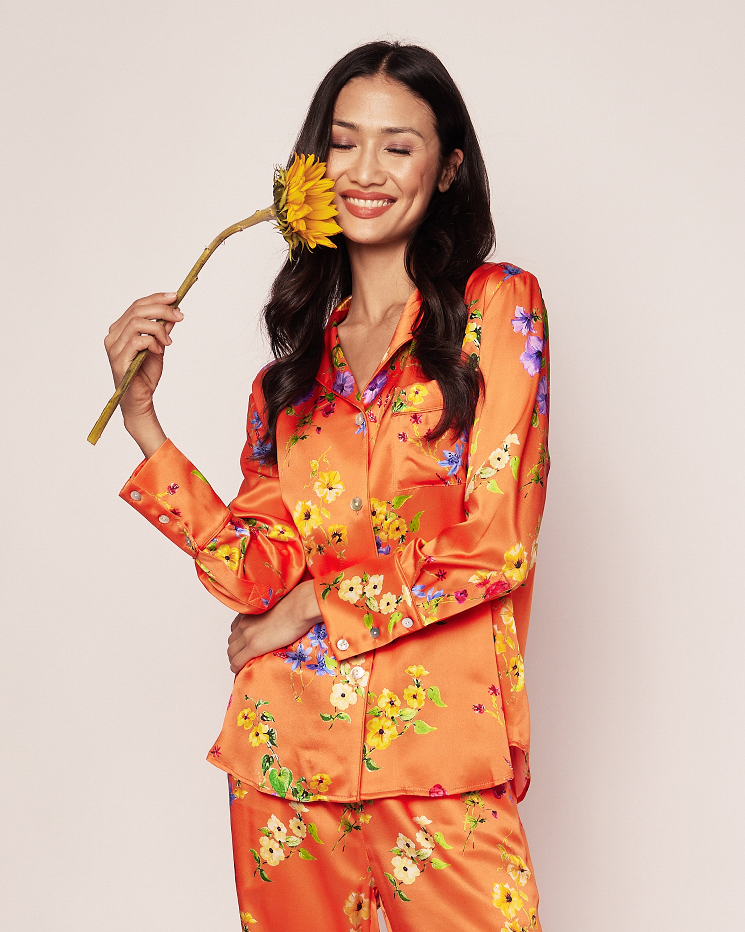 A person smiles while holding a sunflower, dressed in Petite Plumes Womens Silk Pajama Set in Tangerine Brilliant Botanical against a plain backdrop, exuding elegance.