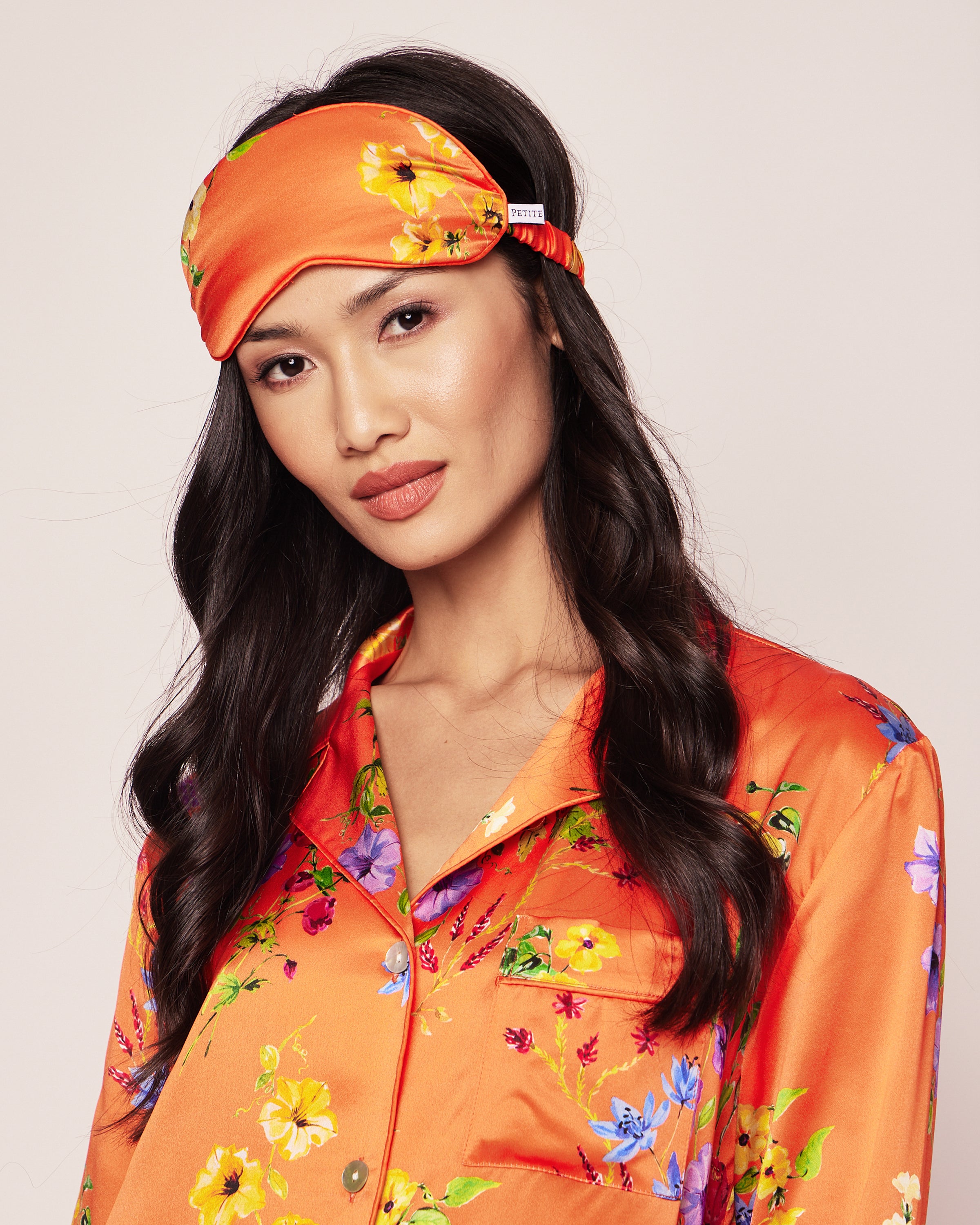 A person poses against a plain background, wearing Petite Plumes Womens Silk Sleep Mask in Tangerine Brilliant Botanical and matching pajamas, with long dark hair.