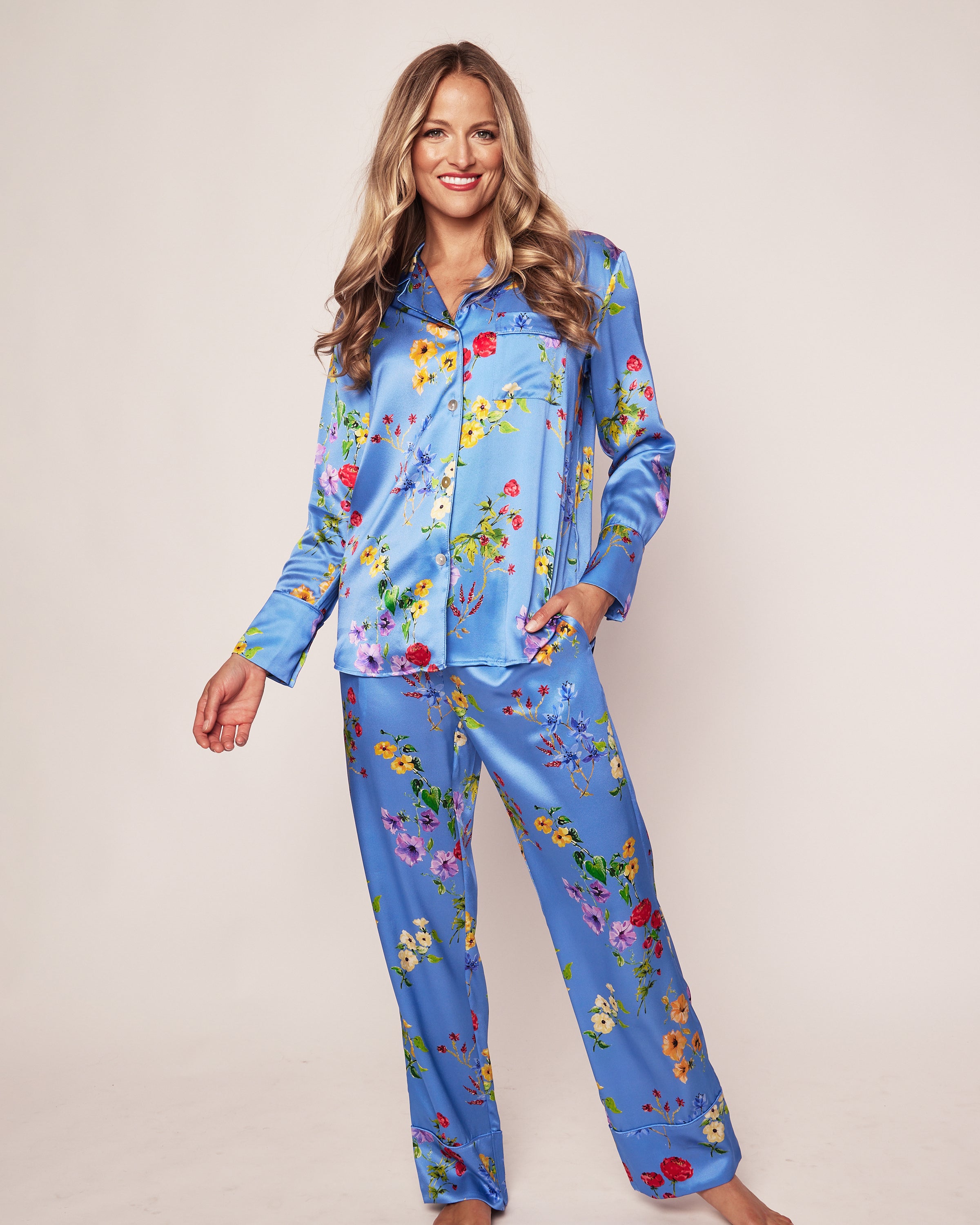 A person with long hair smiles, wearing Petite Plumes Womens Silk Pajama Set in Brilliant Botanical Azure. The luxuriant floral pattern contrasts beautifully against a neutral backdrop, enhancing the scene with elegance and charm.