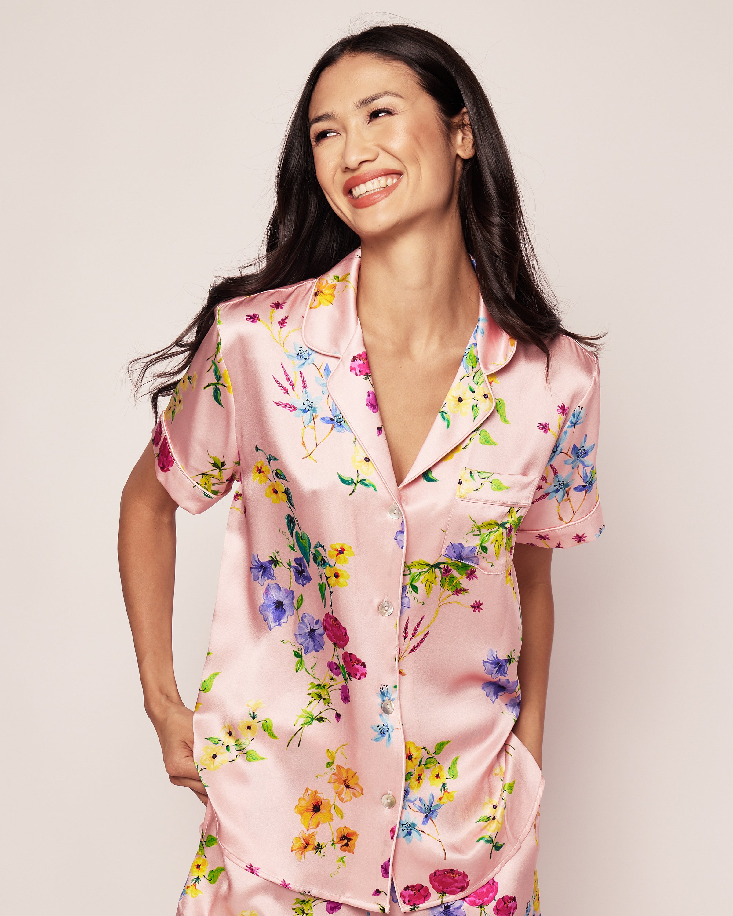 A person smiling and looking to the side wearing the Petite Plume Womens Silk Pajama Short Set in Blush Brilliant Botanical, featuring colorful flowers with a collared neckline and button closure against a plain background.