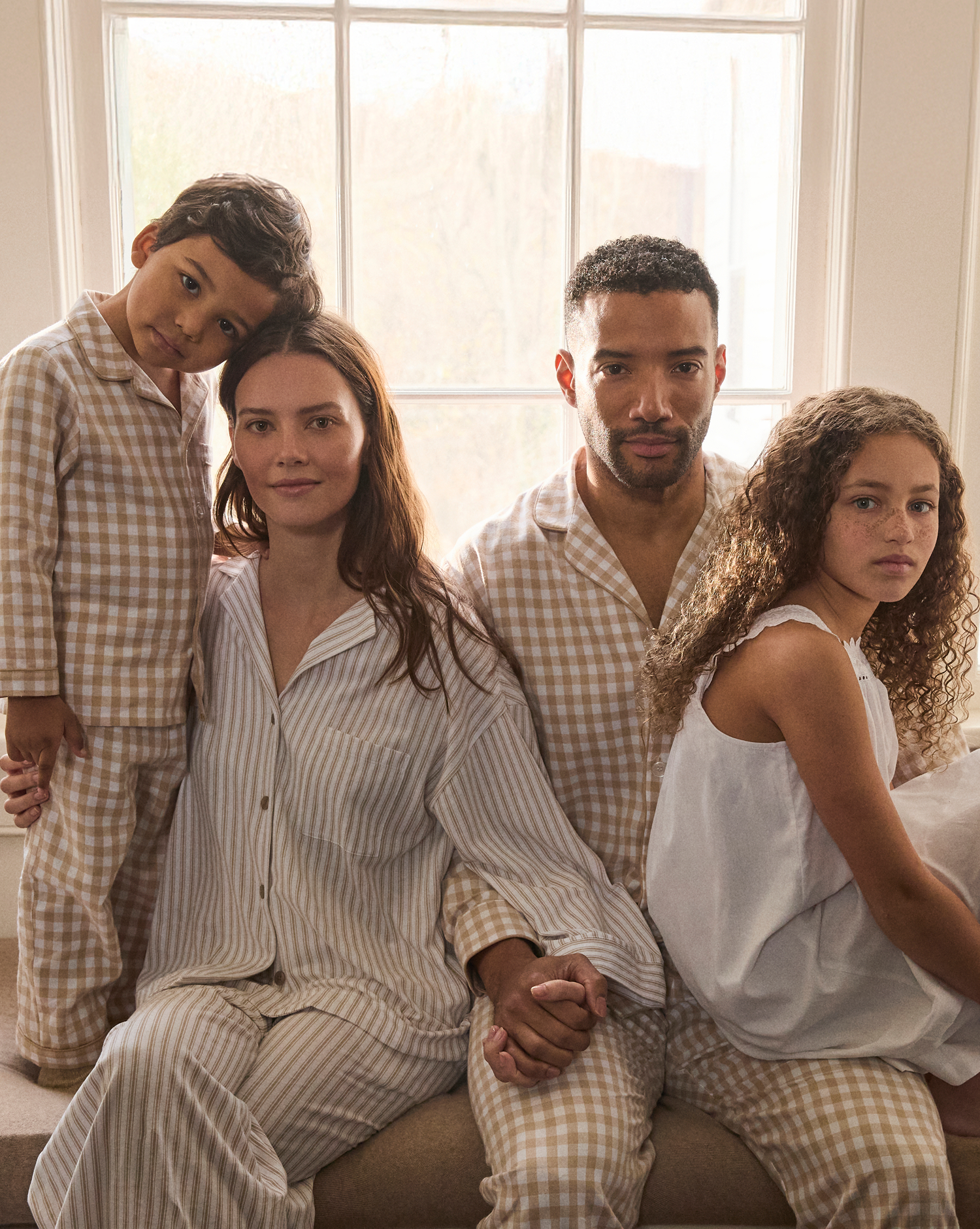 A family of four sits together by a bright window, all wearing neutral-toned pajamas. The parents hold hands, with their young son leaning on his mother and their daughter sitting beside her father.