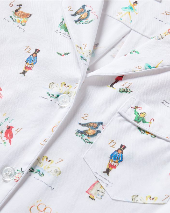 White pajama shirt with colorful illustrations and numbers inspired by The Twelve Days of Christmas, including birds, dancers, and drummers. Shirt has white buttons and a front pocket.