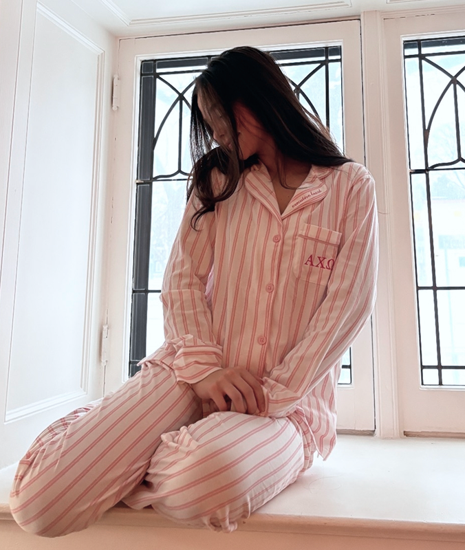 A person wearing pink-and-white striped pajamas sits cross-legged on a windowsill, looking down with their hair falling over their face. Sunlight streams through the tall, decorative windows behind them.
