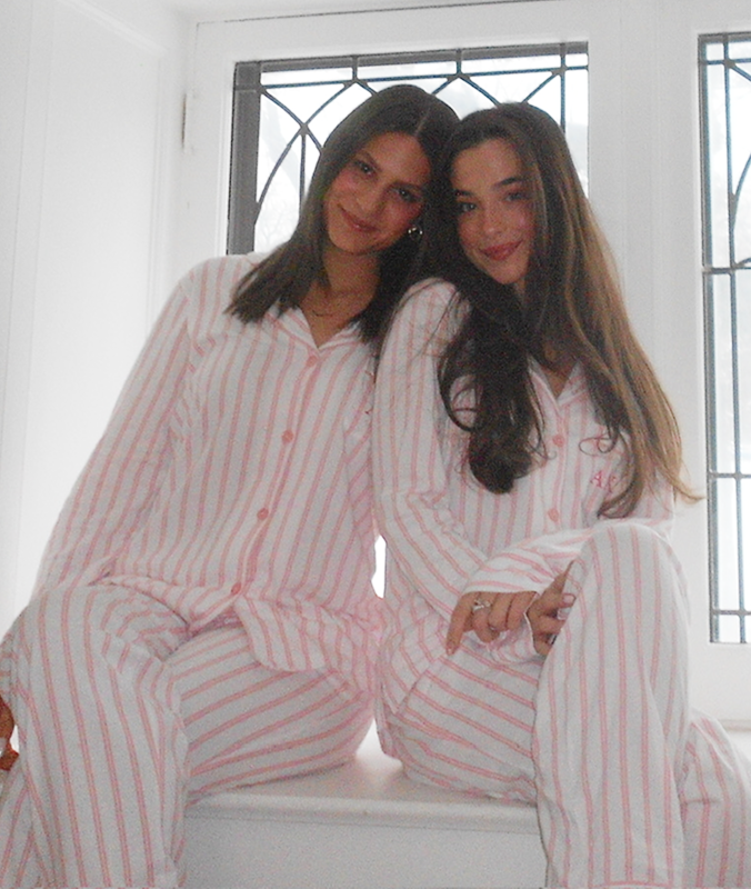Two women wearing matching white pajamas with pink stripes sit side by side on a windowsill, smiling at the camera. A window with decorative black trim is behind them.