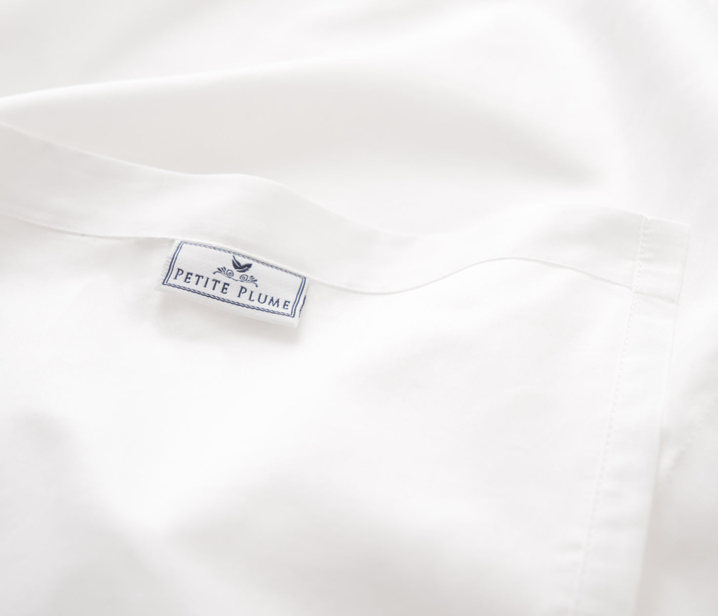 A close-up of a white fabric with a sewn-in label that reads PETITE PLUME in blue letters, featuring a small feather icon above the text. The fabric appears smooth and soft.