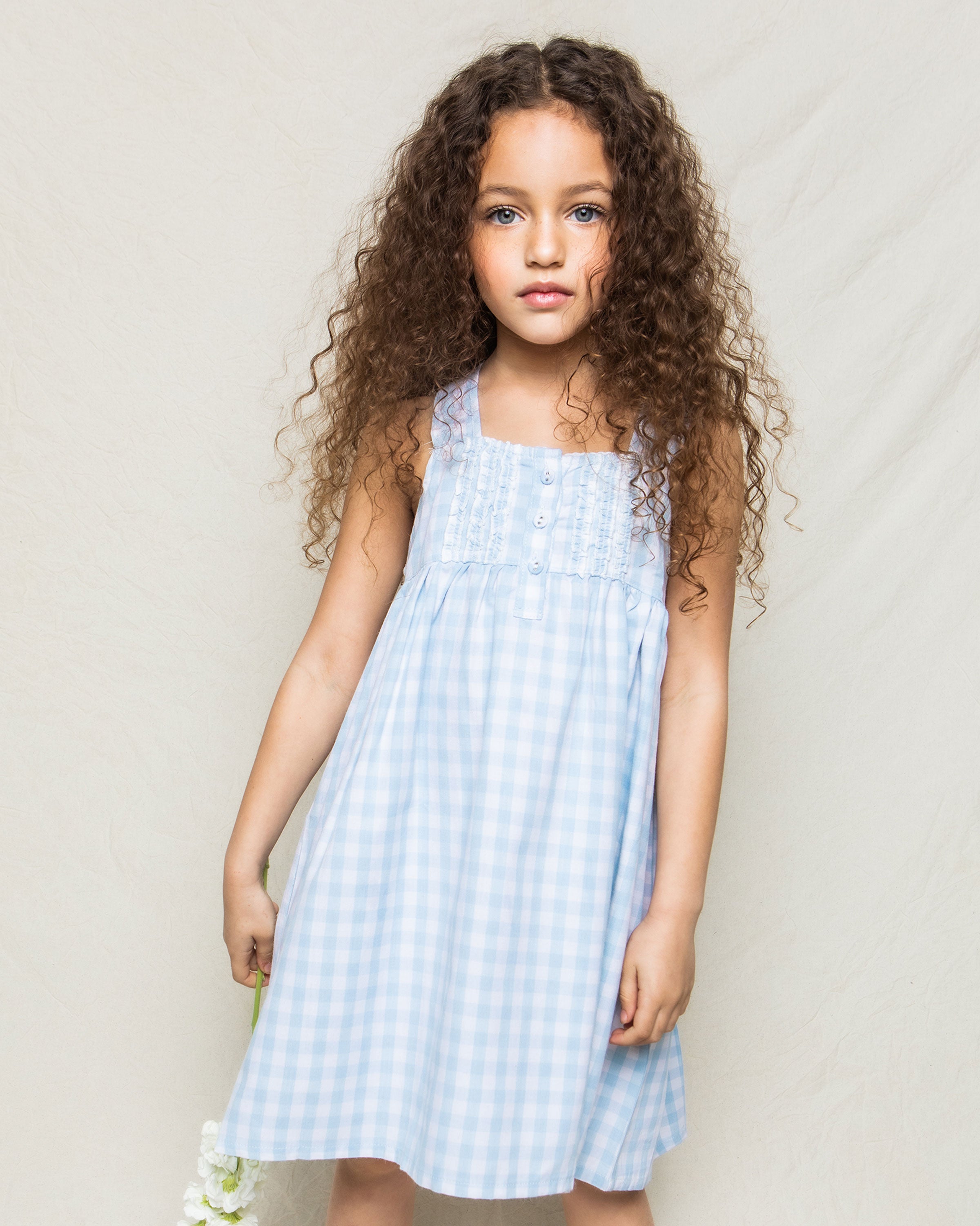 A young girl with long curly hair, wearing a Petite Plume Girls Twill Charlotte Nightgown in light blue gingham, stands against a light background, holding white flowers.