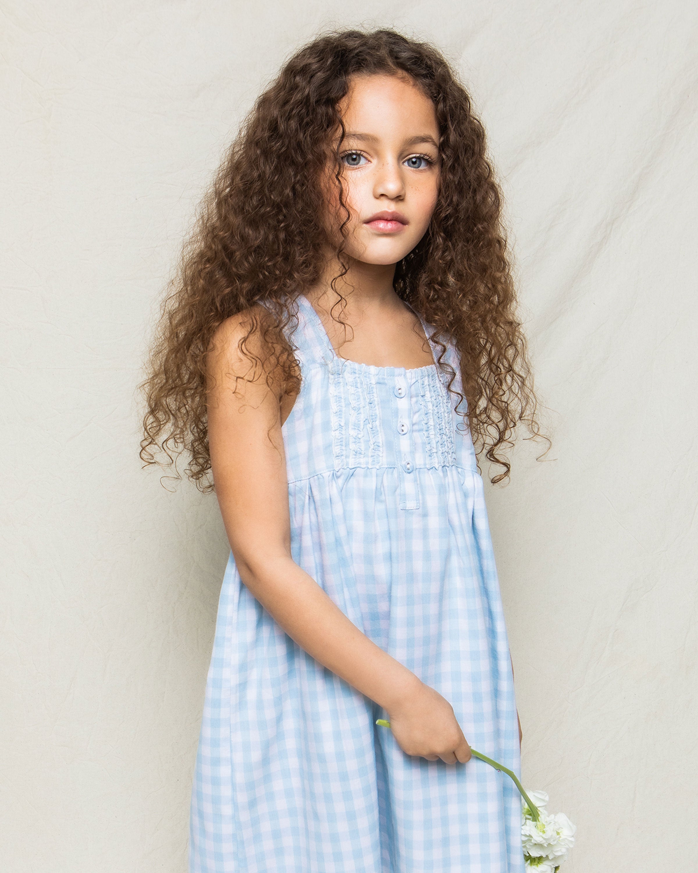 A young girl with long curly hair wears the Petite Plume Girls Twill Charlotte Nightgown in light blue gingham, holding a white flower. She stands against a neutral background, looking towards the camera.