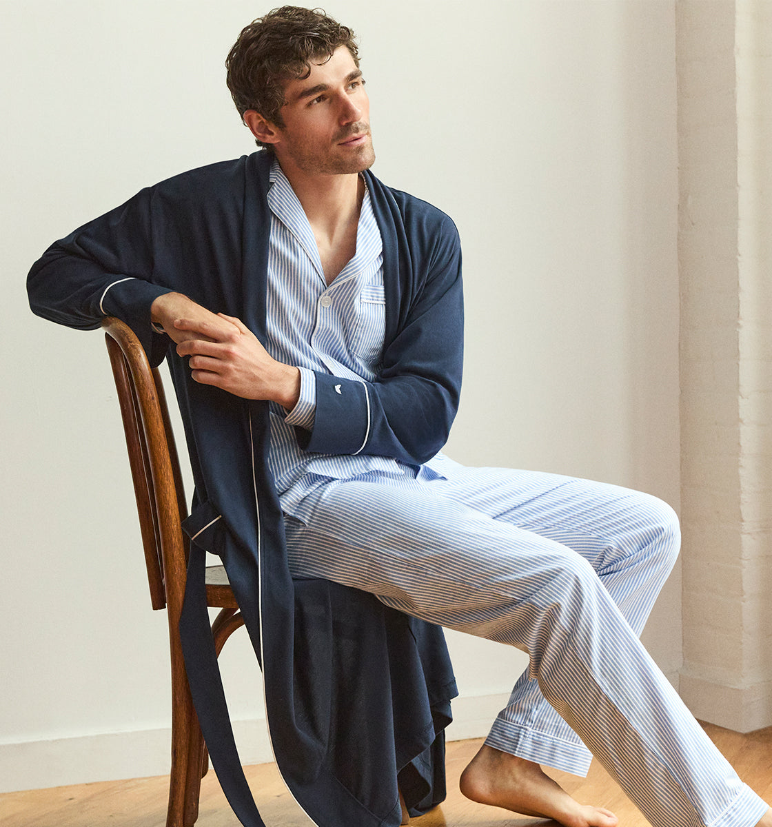 A man wearing blue and white striped pajamas with a navy robe sits sideways on a wooden chair, barefoot, looking thoughtfully to the side in a softly lit room.