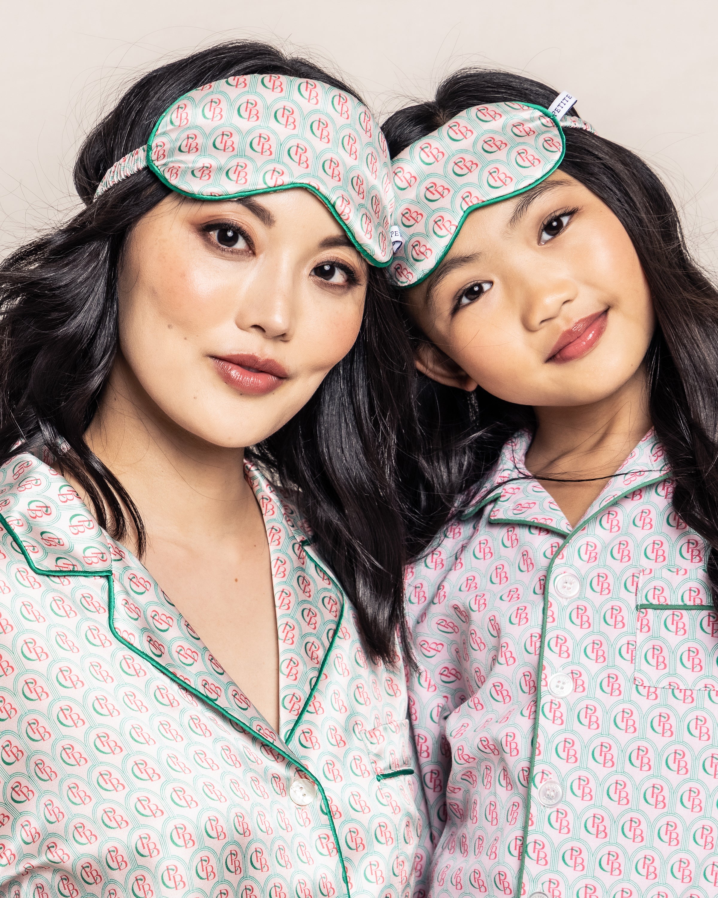 Two people stand close, smiling at the camera against a light beige background. They wear matching pajamas with a pink and green pattern and coordinate with the Colony Hotel x Petite Plume Adults Silk Sleep Mask in Exclusive Print, embodying the promise of the sweetest dreams.