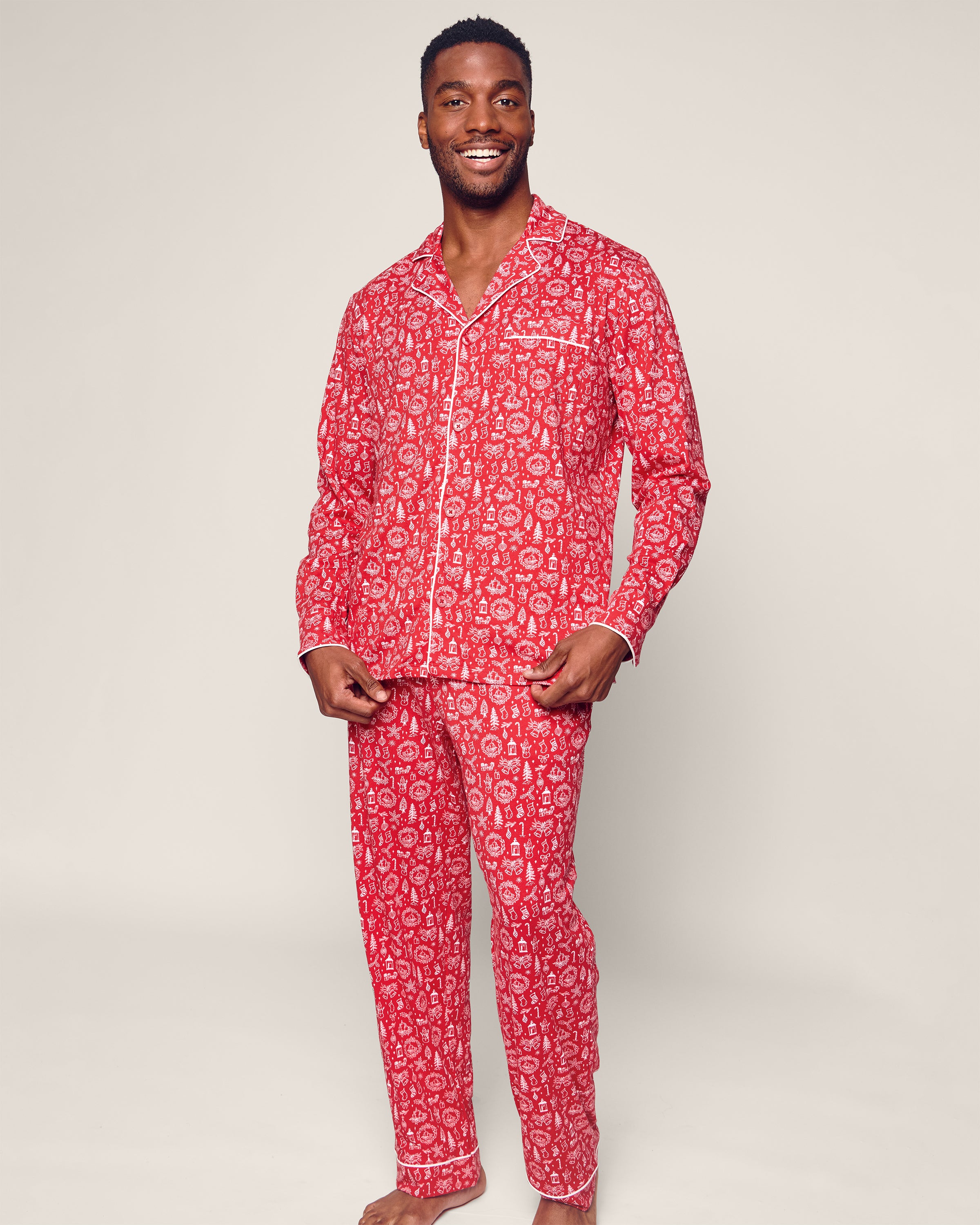 A person stands smiling in a luxurious Mens Pima Pajama Set in Whimsical Winter Saks x Petite Plume, made of Peruvian Pima cotton with a white floral pattern, against an off-white background.