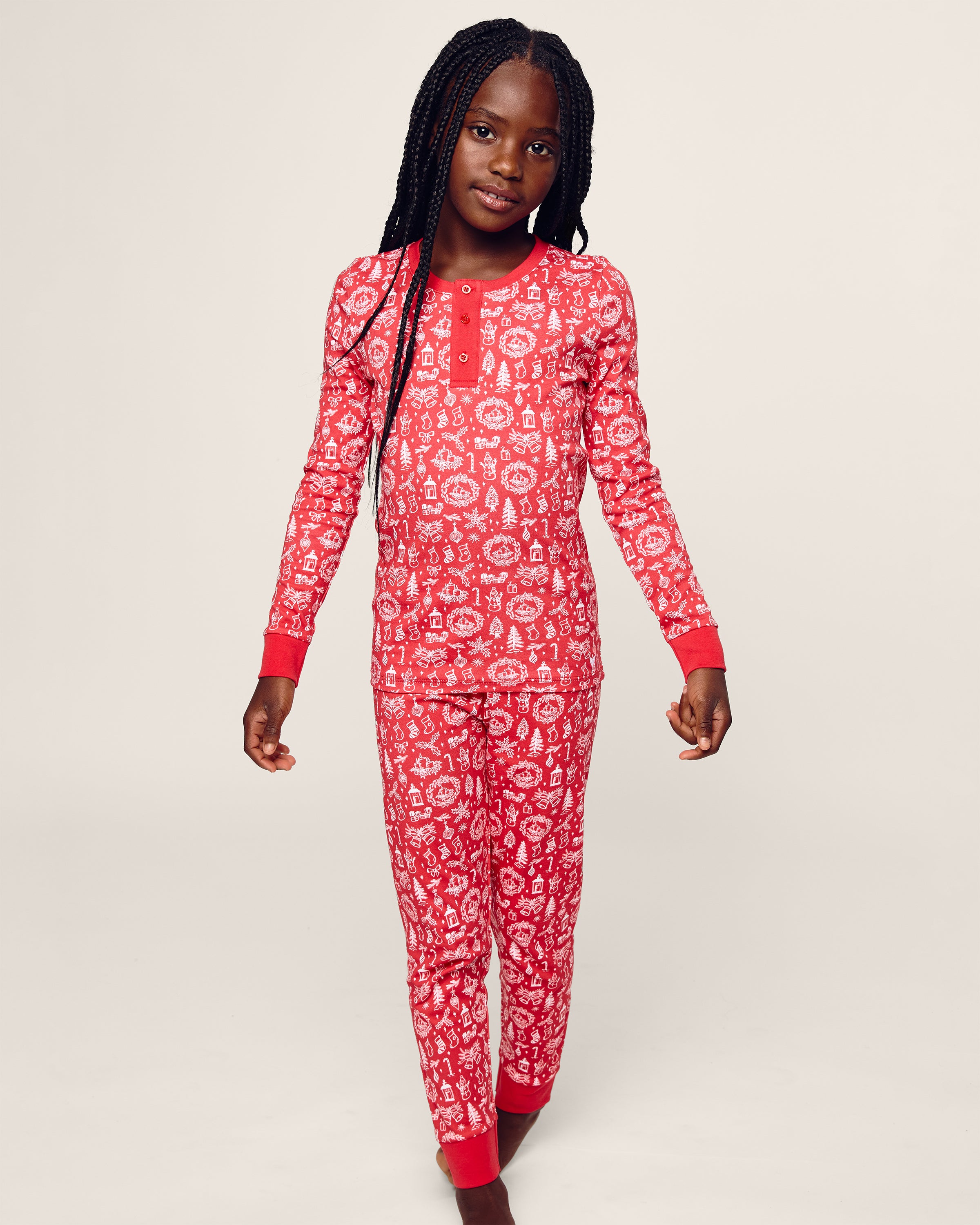 A young girl with long braided hair poses against a plain light background, wearing Petite Plumes Kid’s Pima Snug Fit in Whimsical Winter Saks x PP—red pajamas with a white festive pattern made from cozy Pima cotton. The sleepwear includes a long sleeve top with cuffs at the wrists and ankles.