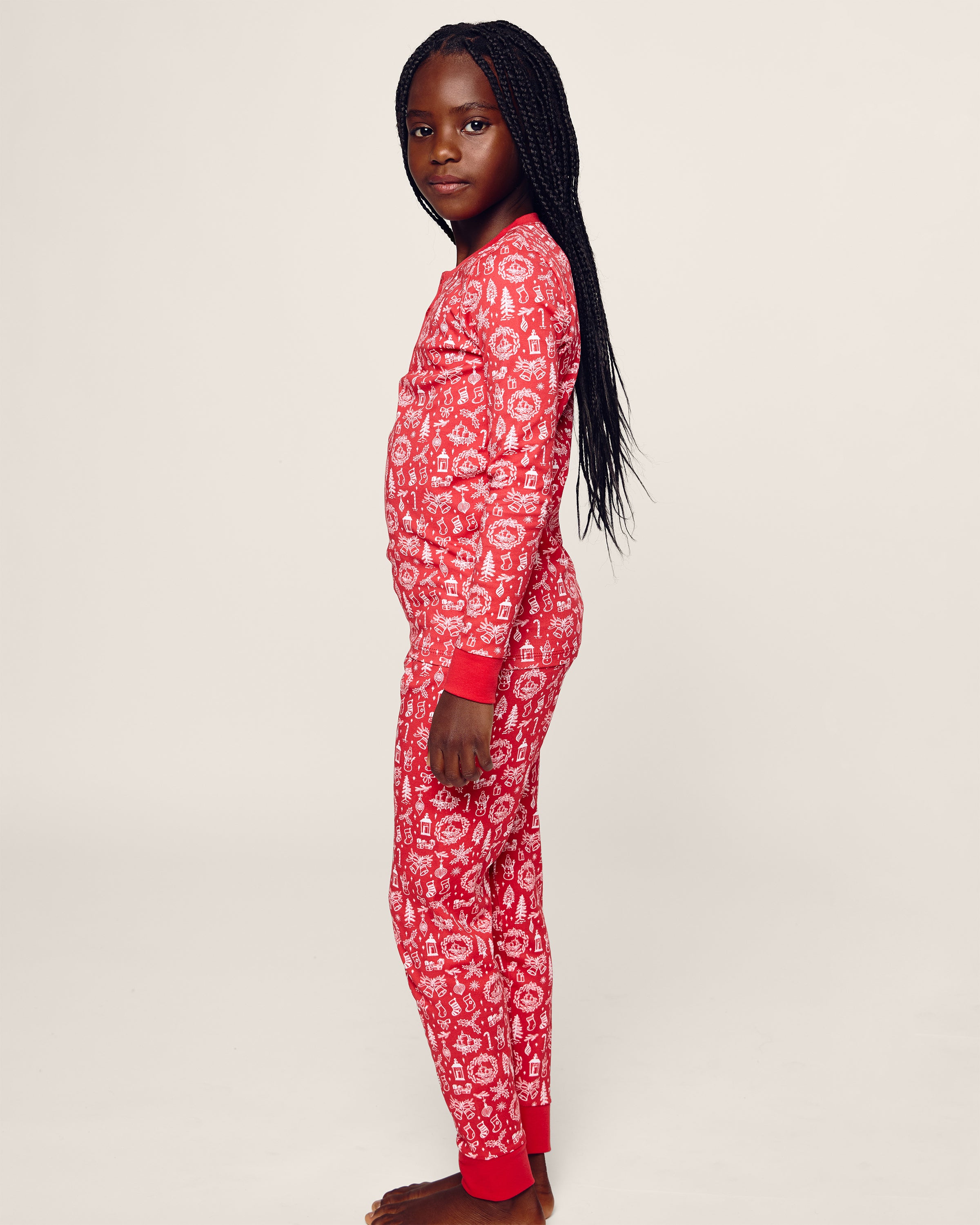 A young girl stands barefoot against a plain background, wearing Petite Plumes Kids Pima Snug Fit in Whimsical Winter Saks x PP sleepwear. The red pajamas made from soft Pima cotton feature a long sleeve top with white patterns. She faces slightly right with a relaxed expression.