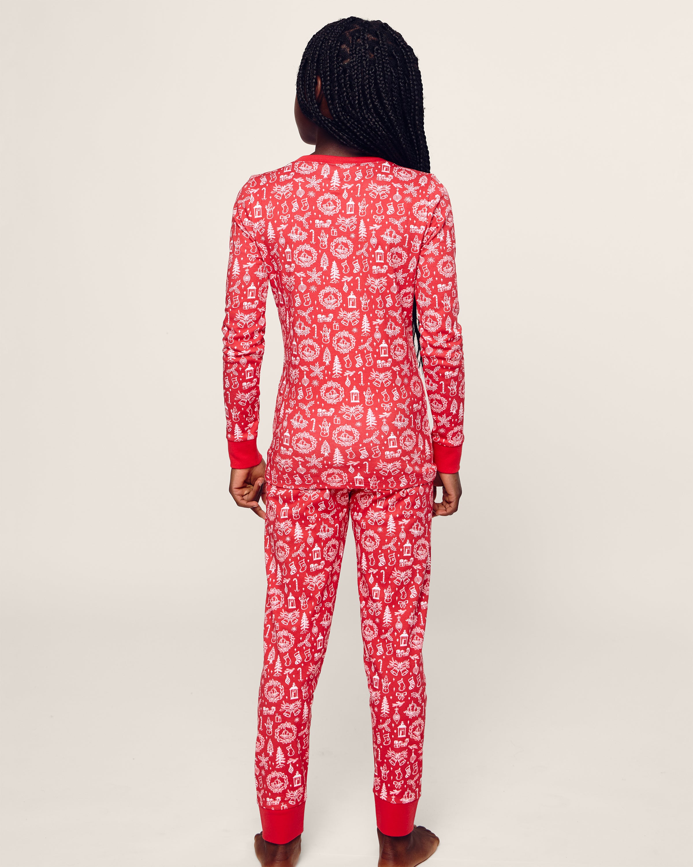 A child with braided hair faces away, wearing the Kids Pima Snug Fit in Whimsical Winter Saks x PP onesie by Petite Plume. Crafted from soft Pima cotton, it features a red background with white holiday snowflakes and reindeer patterns, offering cozy festive charm.