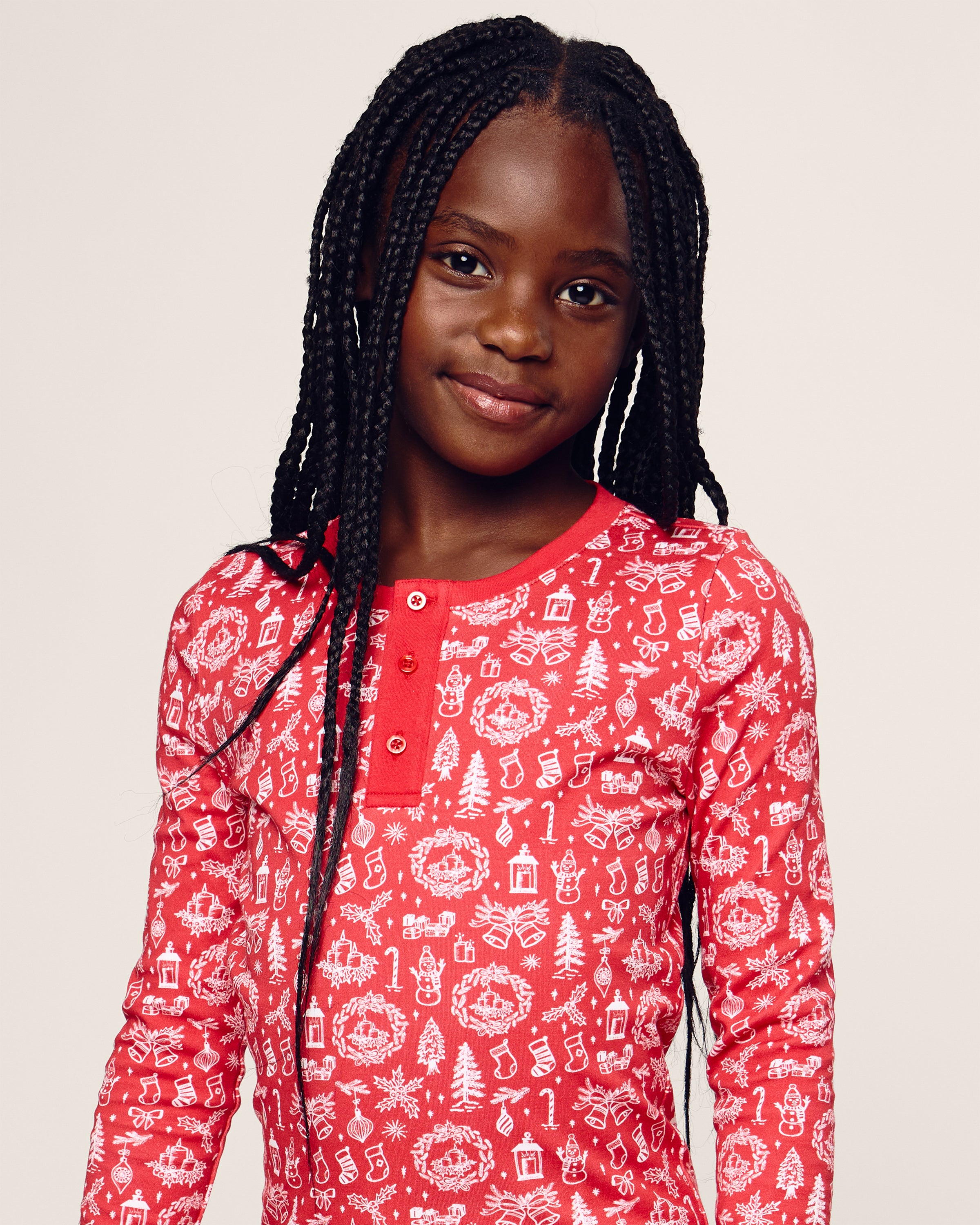 A young girl with long braided hair smiles in a festive red Kids Pima Snug Fit in Whimsical Winter Saks x PP by Petite Plume, featuring white holiday-themed patterns like trees and musical notes, against a plain background.