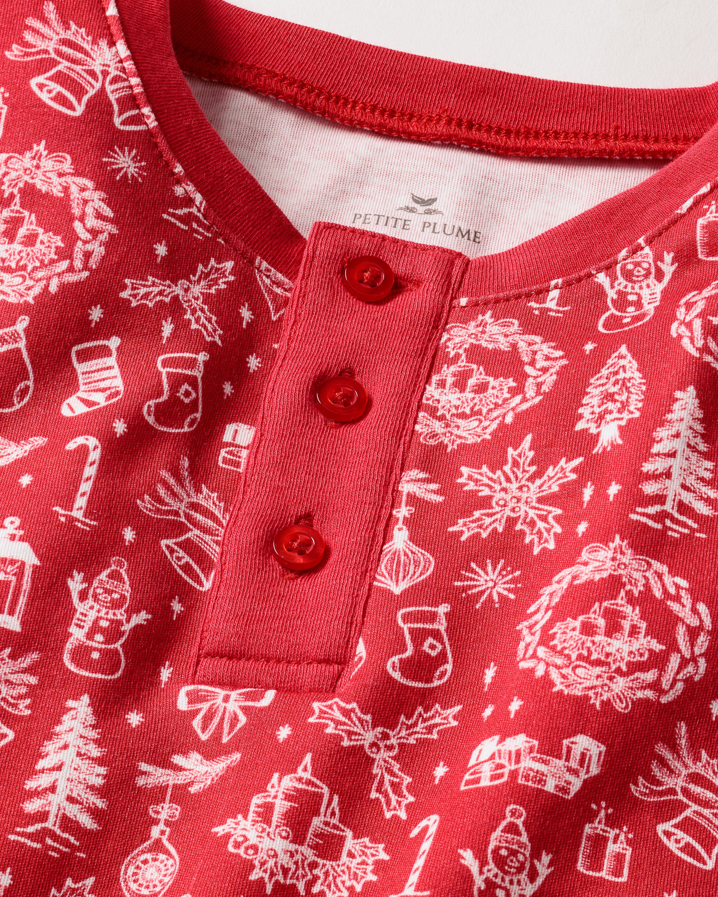 Close-up of the Kids Pima Snug Fit in Whimsical Winter Saks x PP by Petite Plume, a red holiday-themed top made from soft Pima Cotton with white illustrations of snowmen, trees, wreaths, and stockings. It has a three-button placket with red buttons.