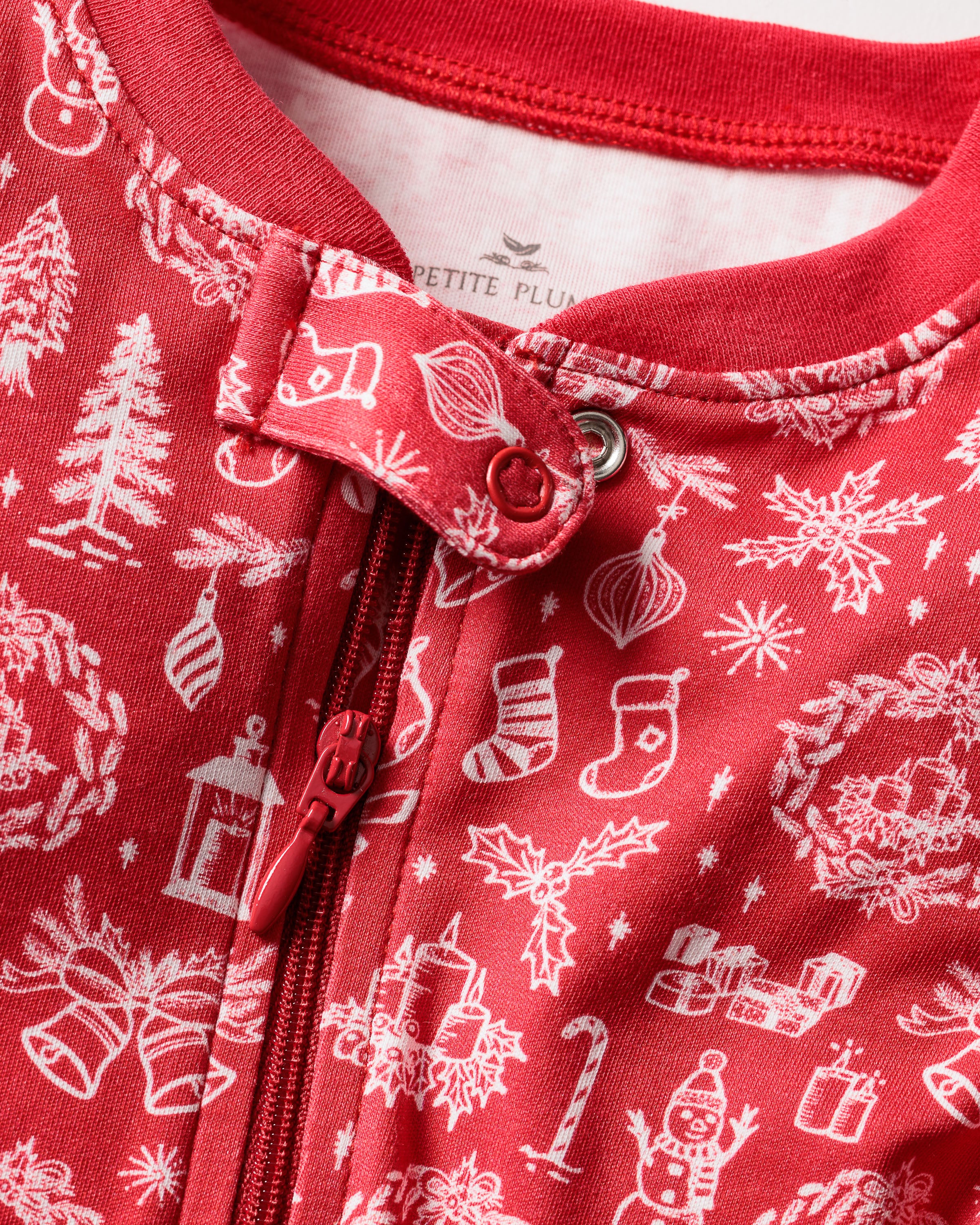 Close-up of Babys Pima Romper by Petite Plume featuring festive holiday patterns like snowmen, trees, stockings, gifts, candy canes, and bells. Made from Pima cotton for softness and durability with a discreet zipper and part of the label visible at the top.