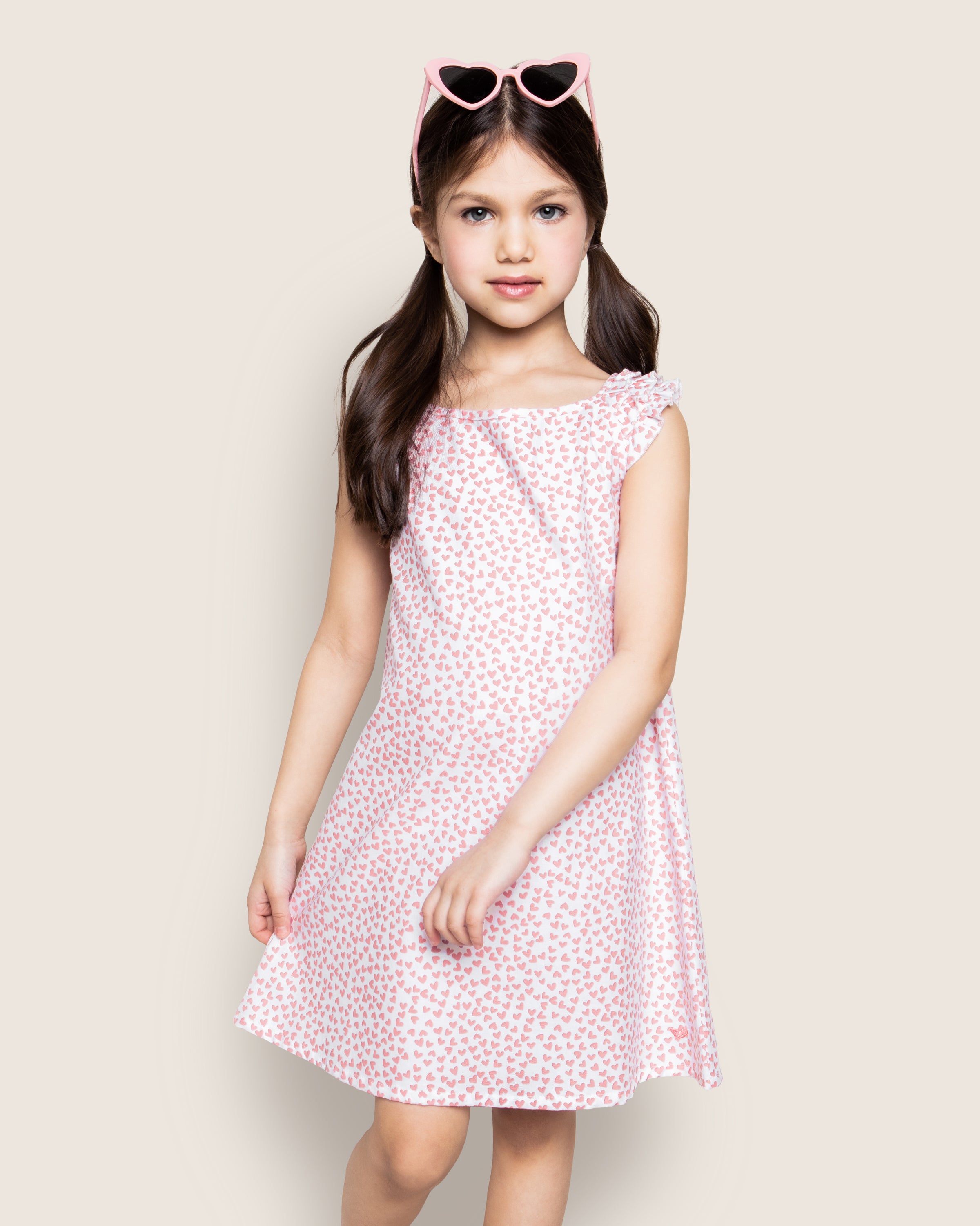 A young girl with long brown pigtails stands wearing the sleeveless Girls Twill Amelie Nightgown in Sweethearts by Petite Plume. She sports heart-shaped sunglasses on her head, set against a plain, light background.