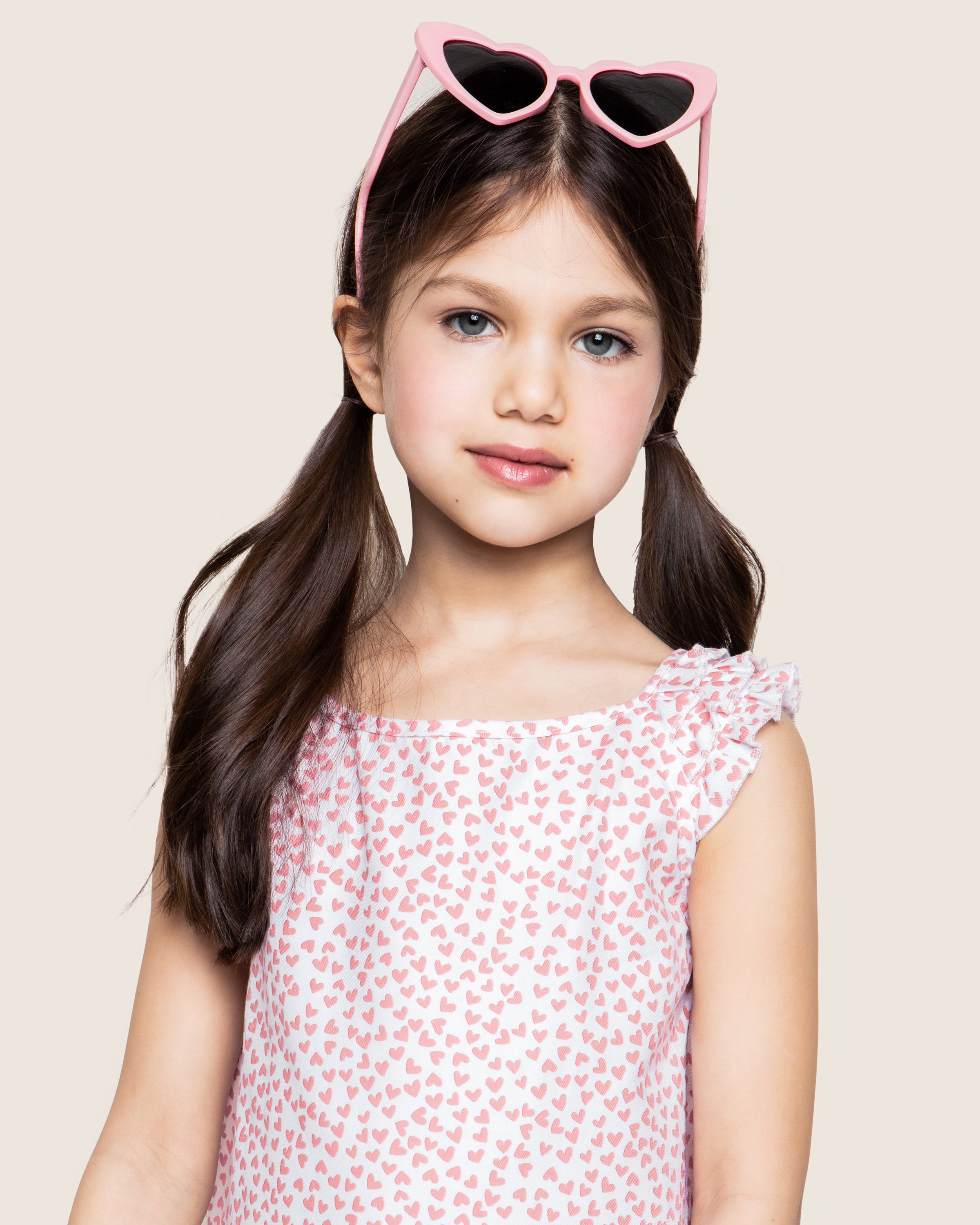 A young girl with long brown hair and pigtails wears Petite Plumes Girls Twill Amelie Nightgown in Sweethearts. She smiles slightly, her pink heart-shaped sunglasses resting atop her head, against a plain beige background that enhances the serene, luxurious sleepwear vibe.