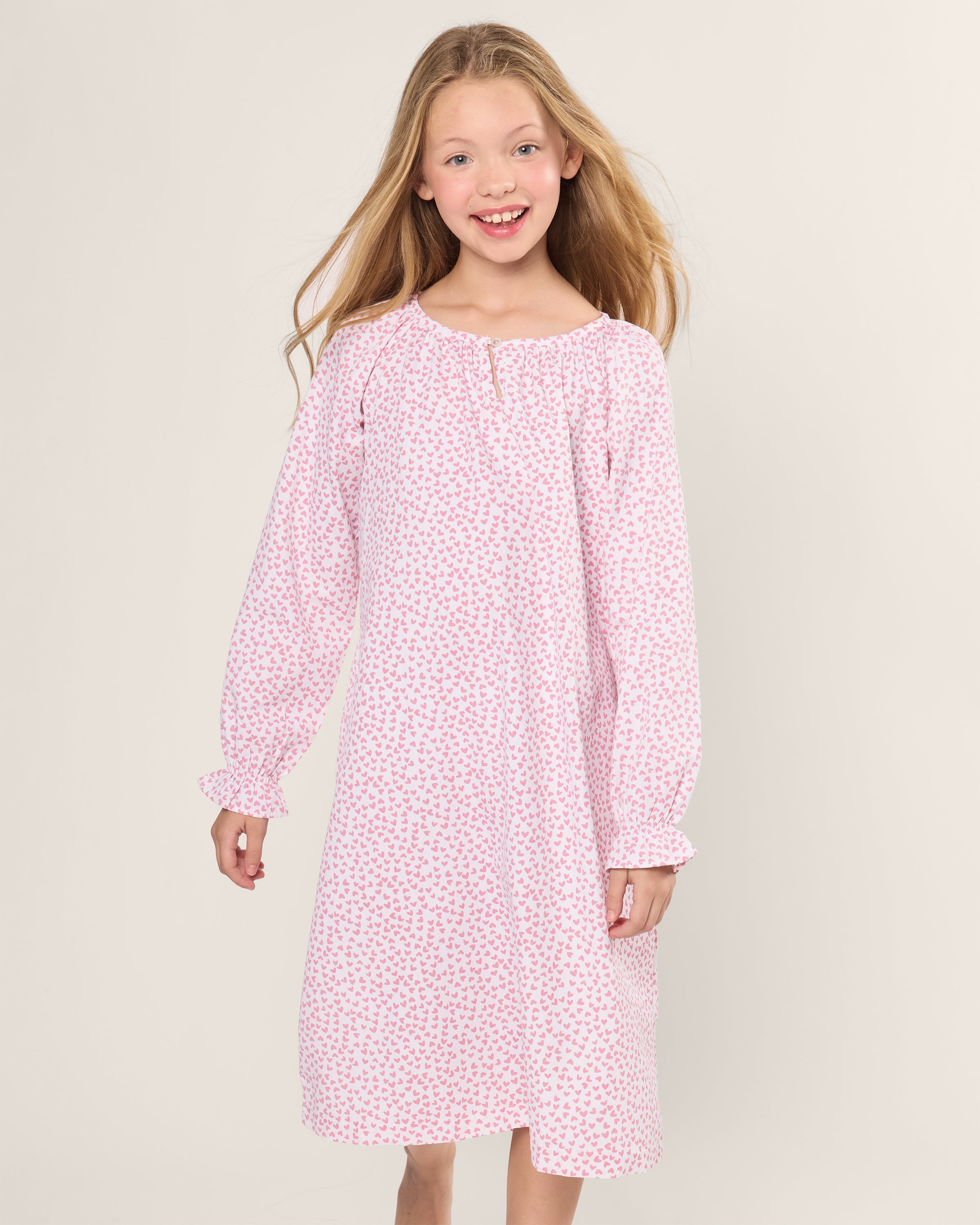 A smiling girl stands wearing Petite Plumes luxurious Girls Twill Delphine Nightgown in Sweethearts. The pink floral design adds charm, and the long-sleeved flame retardant fiber ensures safety and comfort, set against a plain light background.