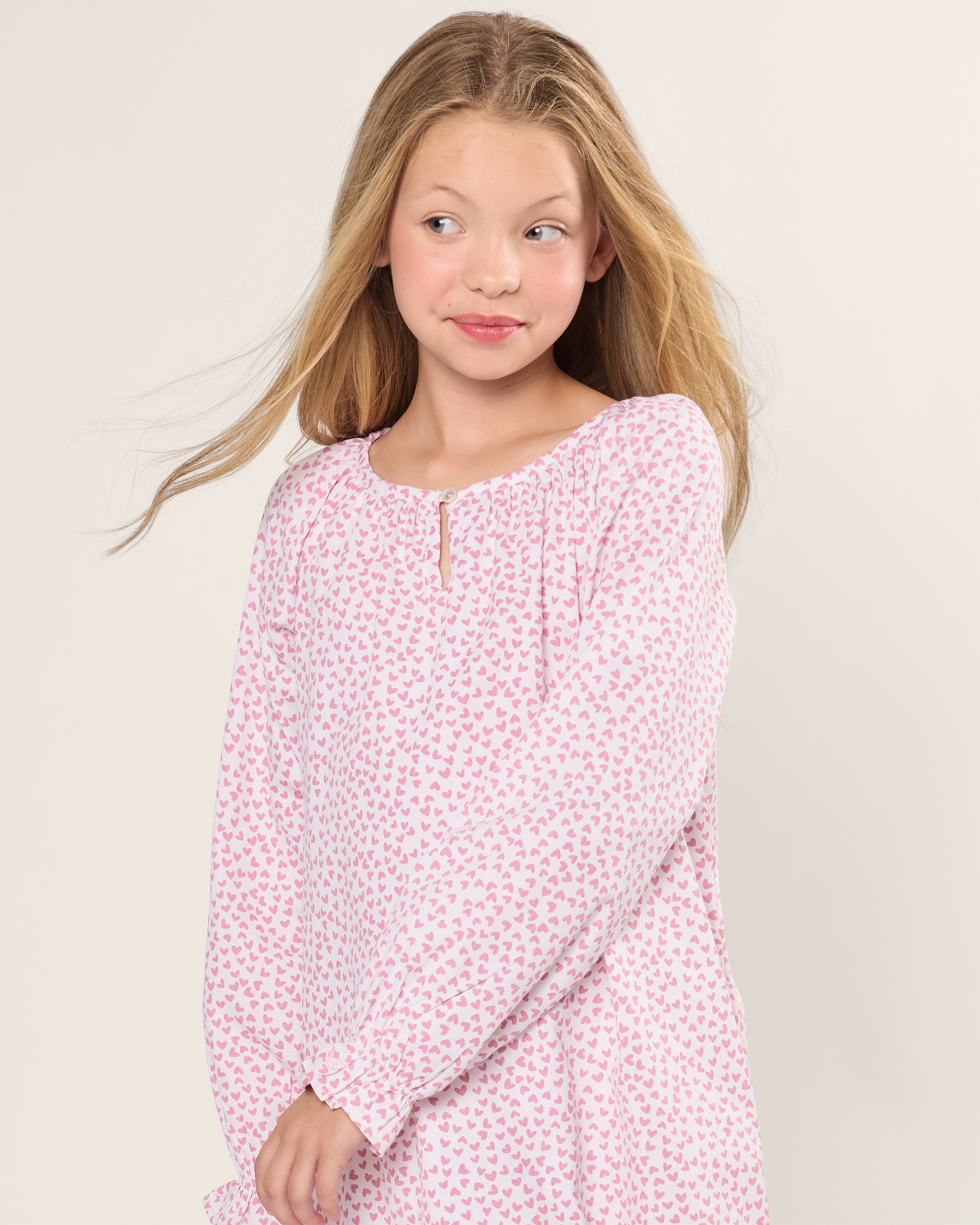 A young girl with long blonde hair stands in a Petite Plume Girls Twill Delphine Nightgown in Sweethearts, its pink and white heart-patterned fabric draping gracefully against a plain background. Her hair partially falls over her shoulder as she gazes to the side with a slight smile.
